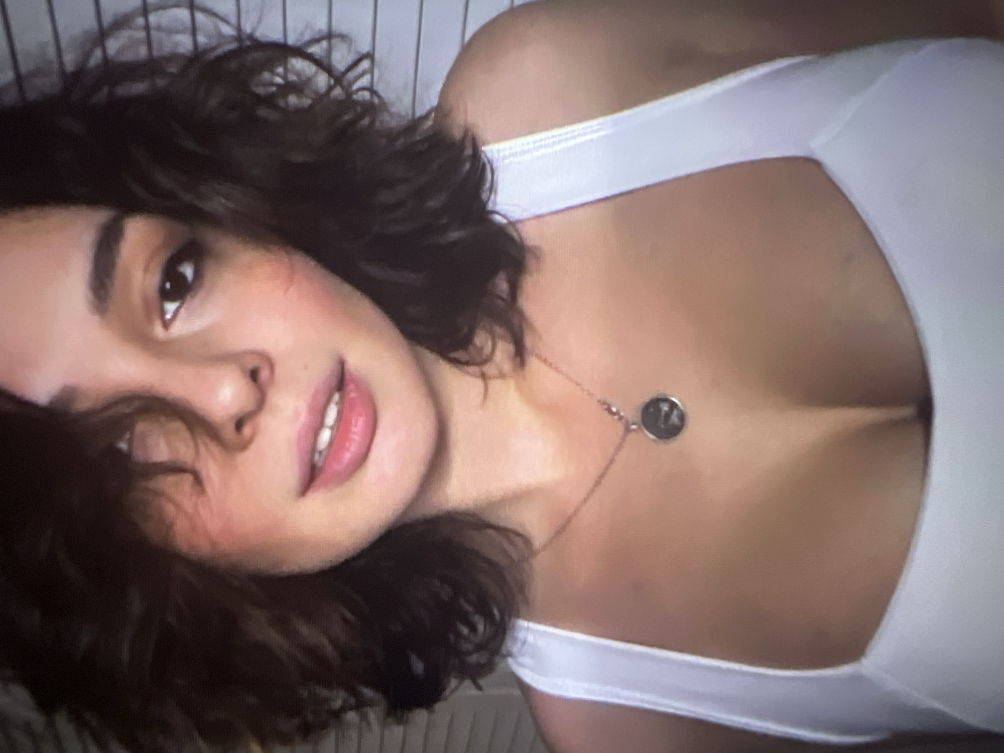 The image is a close-up, slightly blurry portrait of a young woman with dark, curly hair that partially obscures her face. She is looking towards the camera with a soft expression, her lips slightly parted, revealing her teeth. She is wearing a white tank top and a delicate necklace with a circular pendant. The background appears to be a wall with horizontal lines, possibly blinds or paneling. There is no discernible location in the city of Pyatigorsk, Russia.