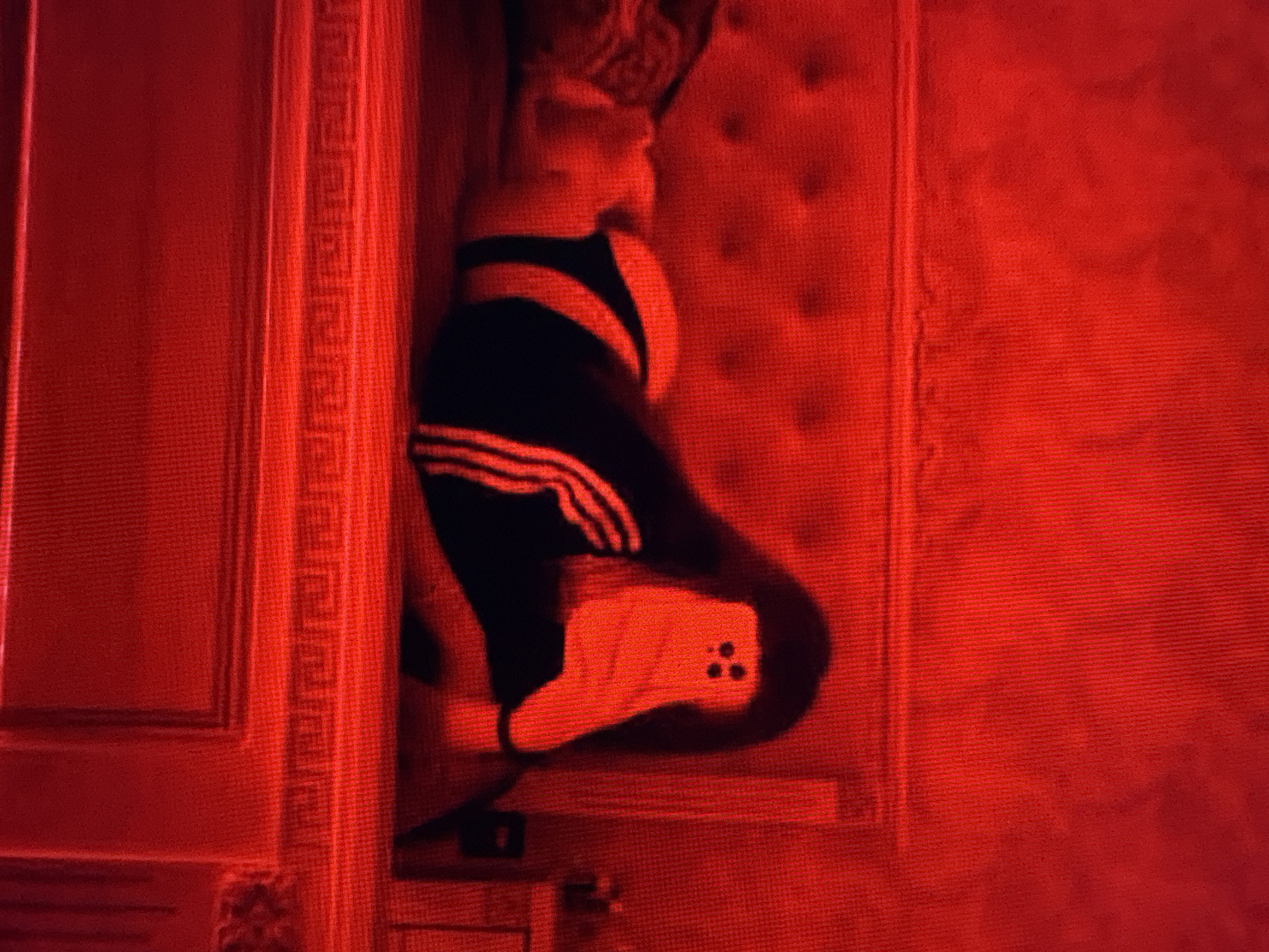 The image shows a person lying on a bed in a room illuminated by red light. The person is wearing black athletic pants with white stripes and a black long-sleeved shirt. They are holding a phone up, presumably to take a selfie, and the phone is obscuring their face. The bed has a tufted headboard. To the right of the bed, a bedside table is partially visible with a small dark object on it. The room appears to be a bedroom. The location is unknown.