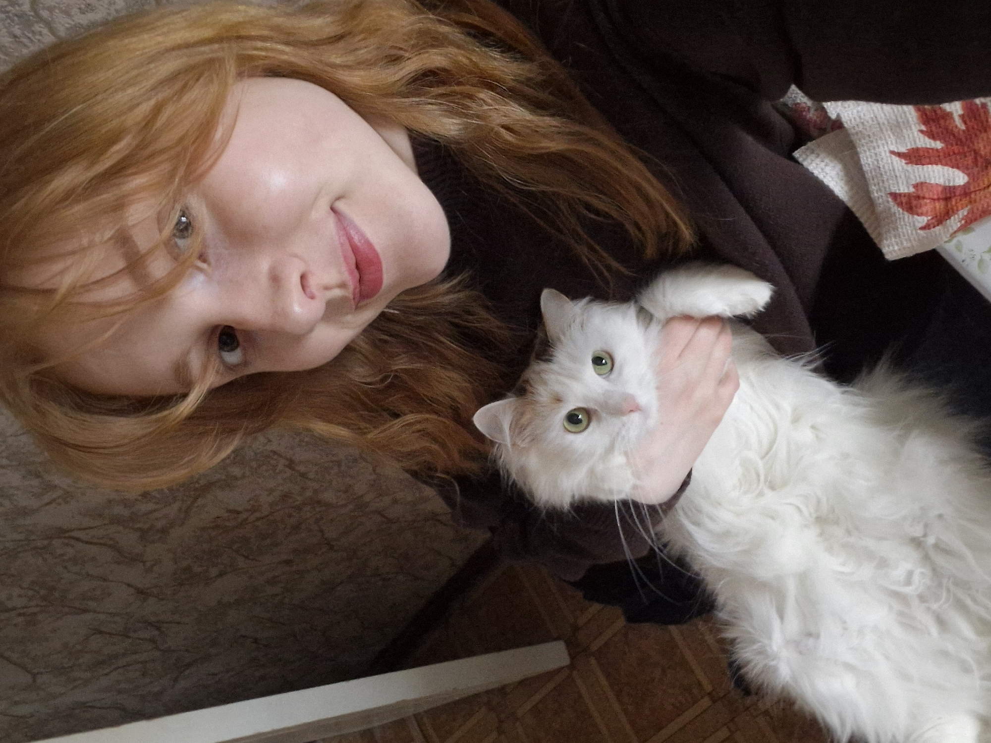This image, taken in Yekaterinburg, Russia, features a young woman and a white cat, likely indoors. The woman, positioned in the upper left portion of the frame, has long, wavy reddish-blonde hair and fair skin. She is smiling and looking directly at the camera, with her lips painted a reddish-pink. She appears to be wearing dark brown clothing, possibly a sweater.
She is holding a fluffy white cat with striking green eyes, which is also looking towards the camera. The cat is cradled in her arm and held gently by her hand. The cat appears calm and comfortable in her embrace.
The setting suggests an indoor environment. Behind the woman's head, a brown, textured surface is visible, possibly a wall or upholstered furniture. To the upper right, near the woman's shoulder, a piece of fabric with a distinct orange and red autumnal leaf pattern can be seen. The lower part of the image shows light brown patterned flooring, possibly tiles or laminate, and a white, linear object, perhaps the edge of furniture or a step, in the bottom left. The overall impression is one of affectionate interaction between the woman and her pet. There is no visible text in the image.