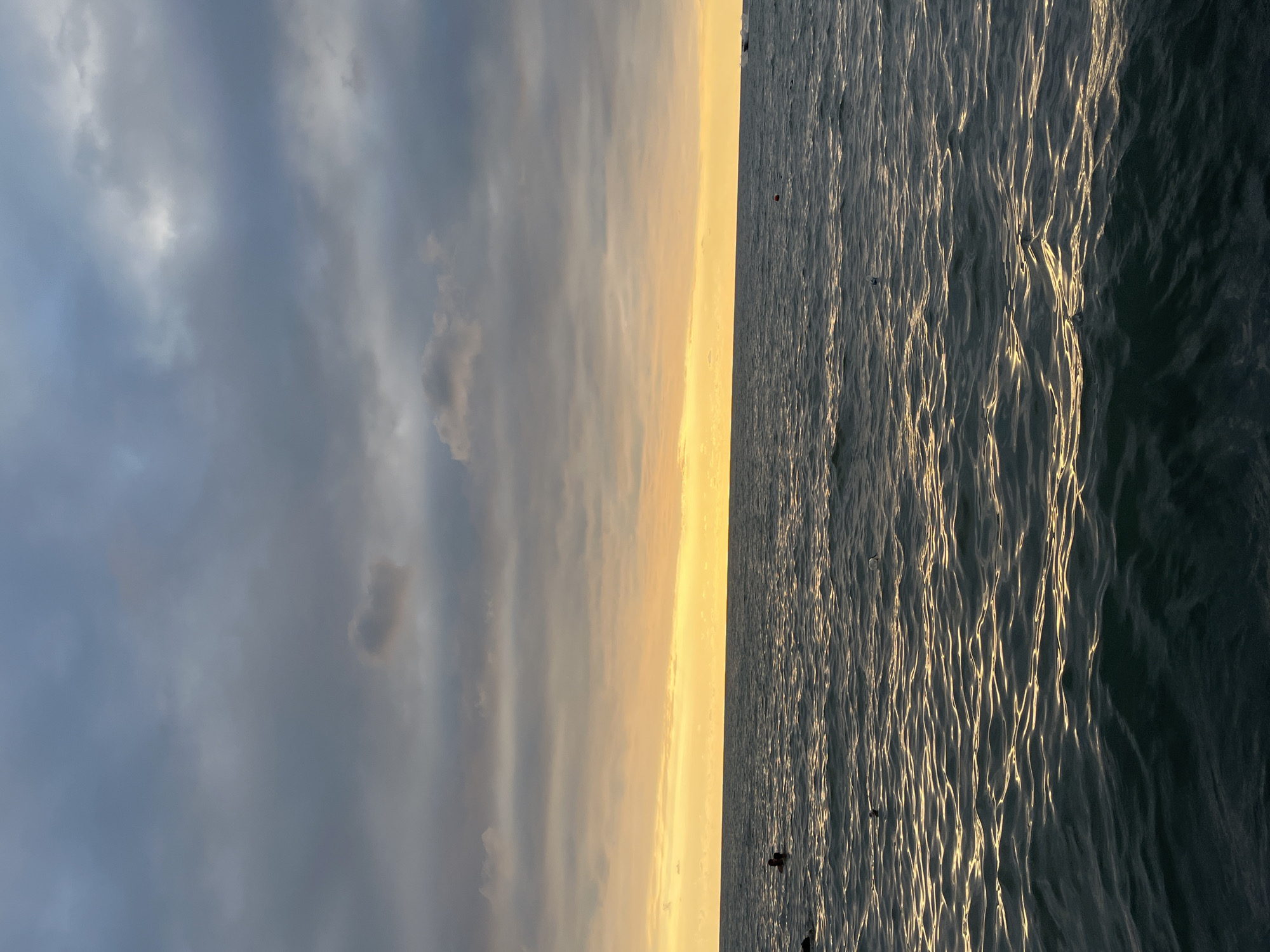 The image depicts a seascape at dusk in Ureki, Georgia. The sky is dominated by heavy, dark grey clouds, with a bright band of yellow and orange light near the horizon, indicating the setting sun. The sea is a deep, dark blue-green, with small, rippling waves that reflect the fading light, creating streaks of golden shimmer. In the distance, on the horizon, a few faint shapes suggest a boat or a distant landmass. In the foreground, there are a few scattered objects on the water, including what appear to be buoys. Two figures are visible in the water on the left side of the image, appearing to be swimming or wading. The overall atmosphere is serene and slightly somber due to the cloudy sky.