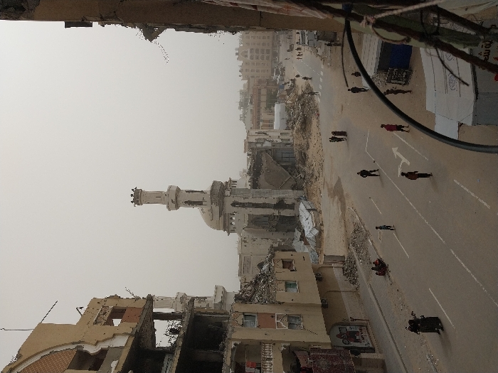 This image depicts a street scene in the city of Khan Yunis, Palestinian Territory, characterized by extensive urban destruction. In the mid-ground, a prominent white mosque with a domed roof and a tall, multi-tiered minaret stands partially damaged, surrounded by rubble. To the left, a multi-story building is heavily destroyed, with large sections of its facade missing, revealing exposed interiors, rebar, and debris. Similarly, a building on the far right edge of the frame also shows severe structural damage.
The central feature is an asphalt road with white lane markings, including a distinctive turning arrow, running through the scene. The areas immediately adjacent to the road are covered in sand and piles of construction debris and rubble.
Approximately 15-20 individuals are present on the street and its periphery. Two women in dark clothing walk together on the left side of the street. A small group of people sits on the ground near the curb on the left. Several individuals, including adults and children in varying attire, are scattered across the roadway, some appearing to be walking, others standing. More people are visible walking in the distance. The sky is overcast and hazy, contributing to a muted, dusty overall appearance of the environment. No vehicles are visible on the street.