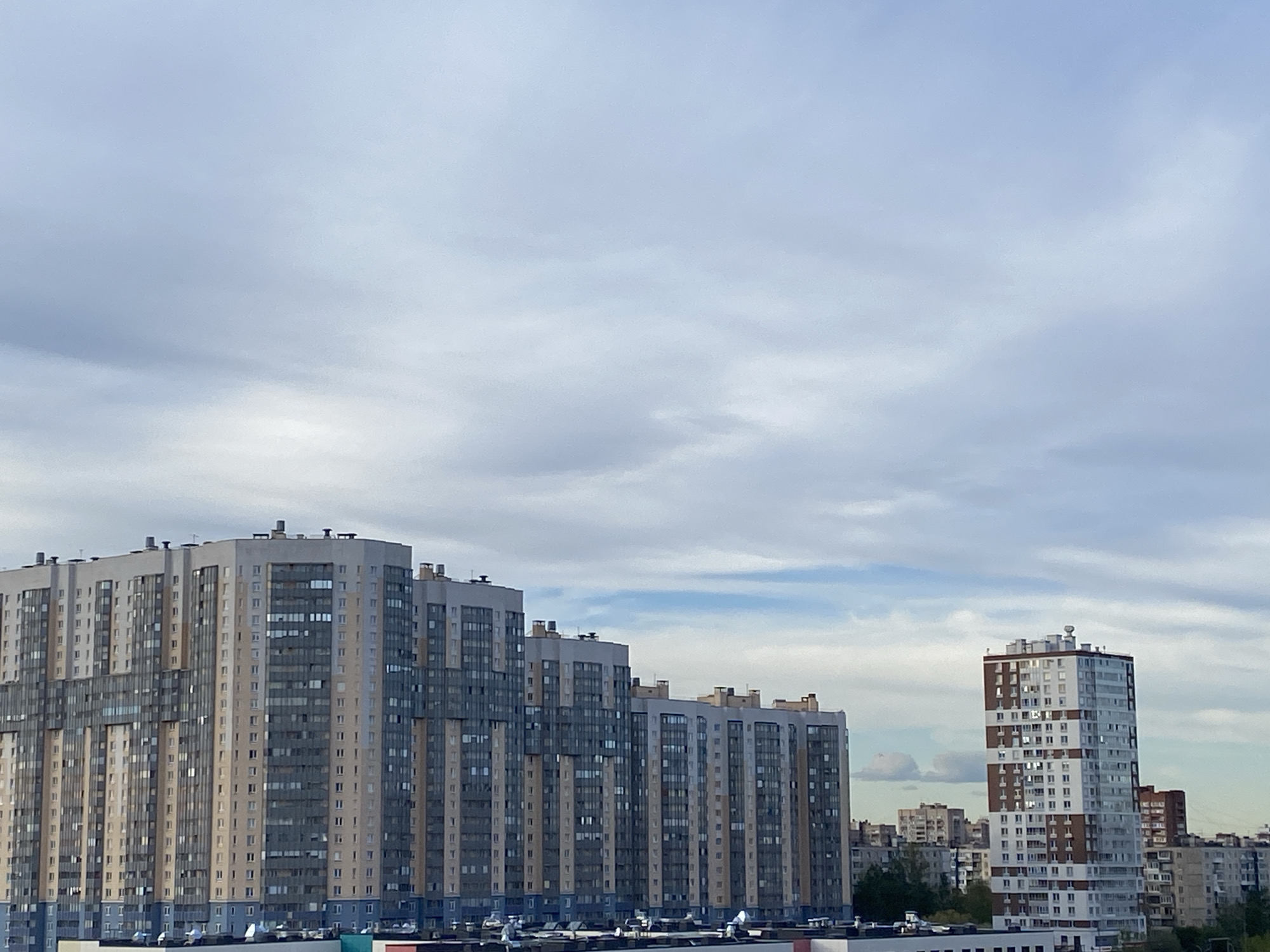 The image depicts a cityscape in Saint Petersburg, Russia, with a focus on large residential buildings under a cloudy sky. The main structures are two large, high-rise apartment complexes. The building on the left is a multi-sectioned structure with varied shades of gray, beige, and blue. The building on the right is a taller, more slender high-rise, with white and brown panels. In the background, a less defined urban skyline is visible, consisting of lower buildings and some greenery. The sky is overcast with varying shades of gray and white clouds. The overall environment suggests an urban setting.
