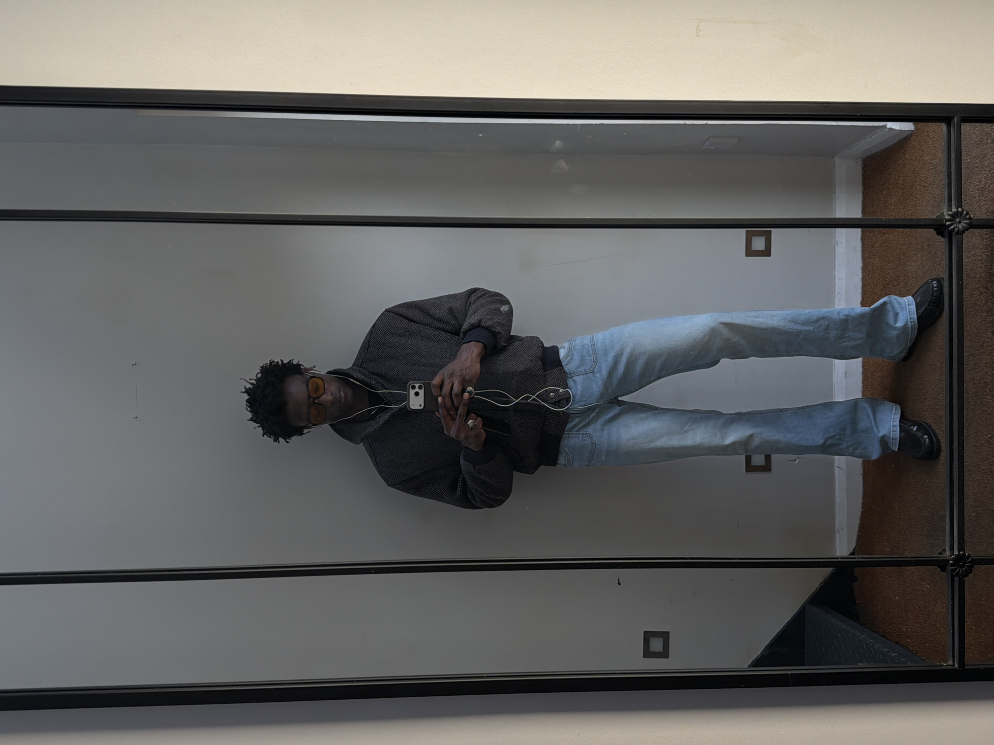 A full-length reflection of a person is visible in a rectangular mirror. The person, a Black male with short, curly hair and yellow-tinted glasses, is wearing a dark textured hoodie, light blue jeans, and black shoes. He is holding a smartphone and wired earphones and appears to be taking a selfie. The reflection also shows a section of a white wall with square electrical outlets and a portion of a brown carpeted area. The mirror itself has a black metal frame with decorative floral elements at the corners. The location is likely inside a building in London, United Kingdom.