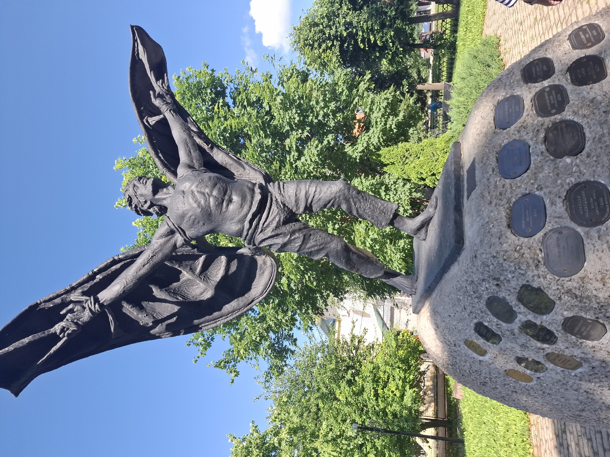 A bronze statue of a winged man is displayed outdoors against a bright blue sky, surrounded by lush green trees. The statue depicts a muscular, semi-nude figure with outstretched arms and large, dark wings. The man's face shows a strained expression, and his body is contorted as if in mid-flight or struggle. He is clad in draped garments that resemble a cape or large fabric billowing behind him.
The statue stands on a rough, light-colored stone base embedded with numerous circular bronze plaques, each bearing visible text. These plaques appear to be commemorative, possibly listing names or significant dates. The base is situated in a park-like setting with neatly mown grass and a paved walkway visible in the background. Sunlight casts shadows, suggesting it is daytime, likely morning or afternoon. The overall mood of the statue is one of drama and perhaps heroism or sacrifice. The location is identified as Kungur, Russia.