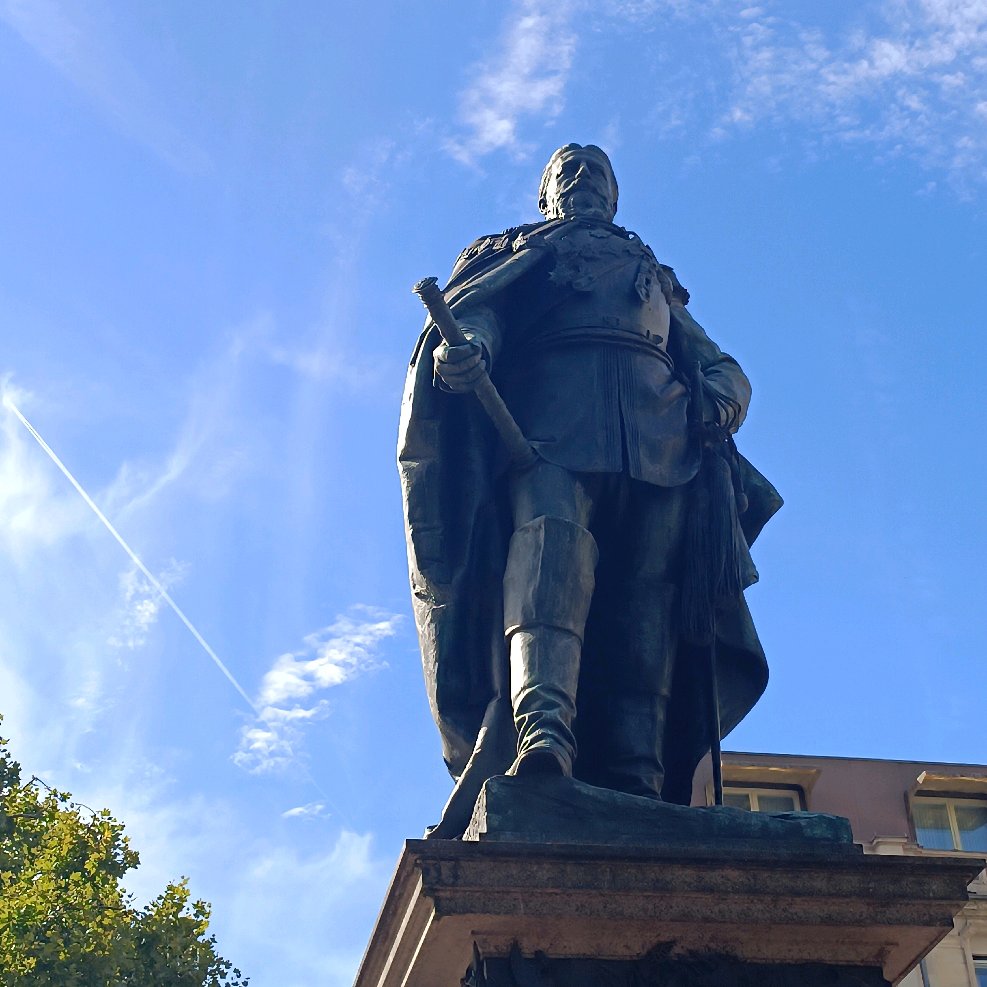This is a low-angle, full-body shot of a large bronze equestrian statue of Kaiser Wilhelm I. The statue is located in the city of Wiesbaden, Germany, specifically on the Kaiser-Friedrich-Platz, in front of the Kurhaus. The statue depicts the Kaiser in military attire, wearing a cape and holding a baton. He is mounted on a horse, which is not fully visible in the frame. The statue stands on a granite pedestal. The background consists of a bright blue sky with some wispy clouds and a faint contrail from an airplane. To the left of the statue, the top of a green tree is visible. In the upper right corner, the roofline of a building with awnings and windows can be seen. The lighting suggests it is daytime, with sunlight illuminating the statue from the side and slightly from above.