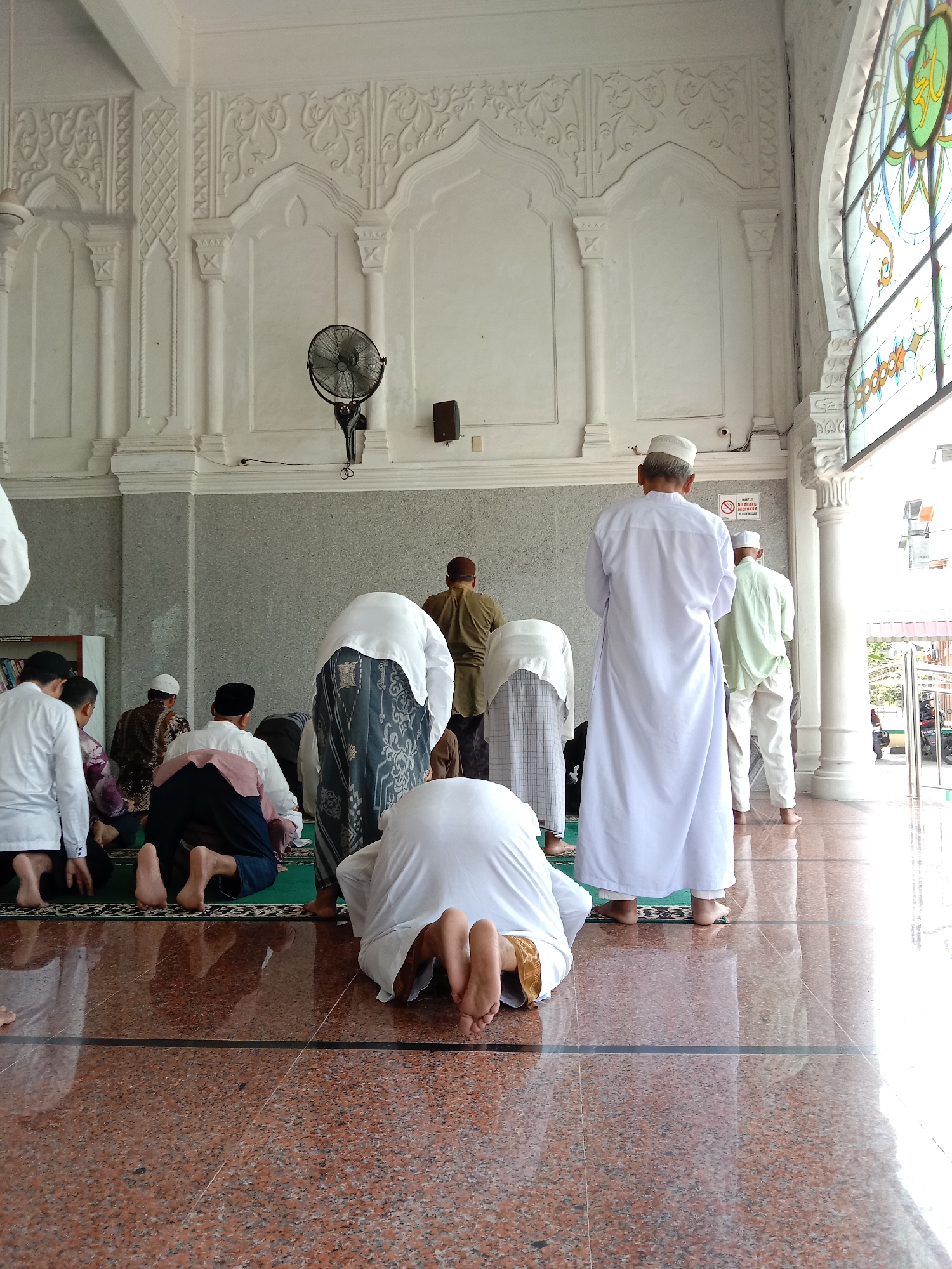 In the city of Lhokseumawe, Indonesia, a group of Muslim men are engaged in prayer inside a mosque. The worshippers are wearing a variety of traditional Islamic clothing, with several donning white robes and head coverings. Some men are kneeling on a green prayer mat, while others are standing or bowing. The mosque's interior features ornate white walls, archways, and a stained-glass window. A wall-mounted fan is visible. The floor is made of polished red marble. A "No Smoking" sign is present. The environment appears to be well-maintained and peaceful.