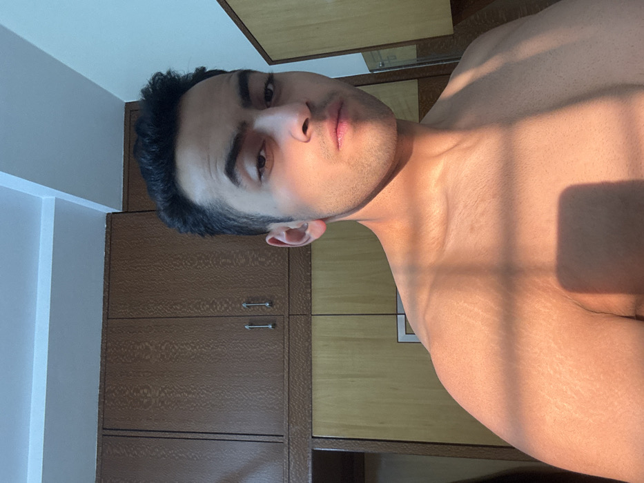 A shirtless man with short, dark hair faces the camera. Sunlight casts shadows across his face and upper body, highlighting his skin. He has dark eyes, a defined jawline, and a slight pout. Behind him, there are wooden cabinets and a wall with a white corner. The image is angled, with the subject tilted to the right.