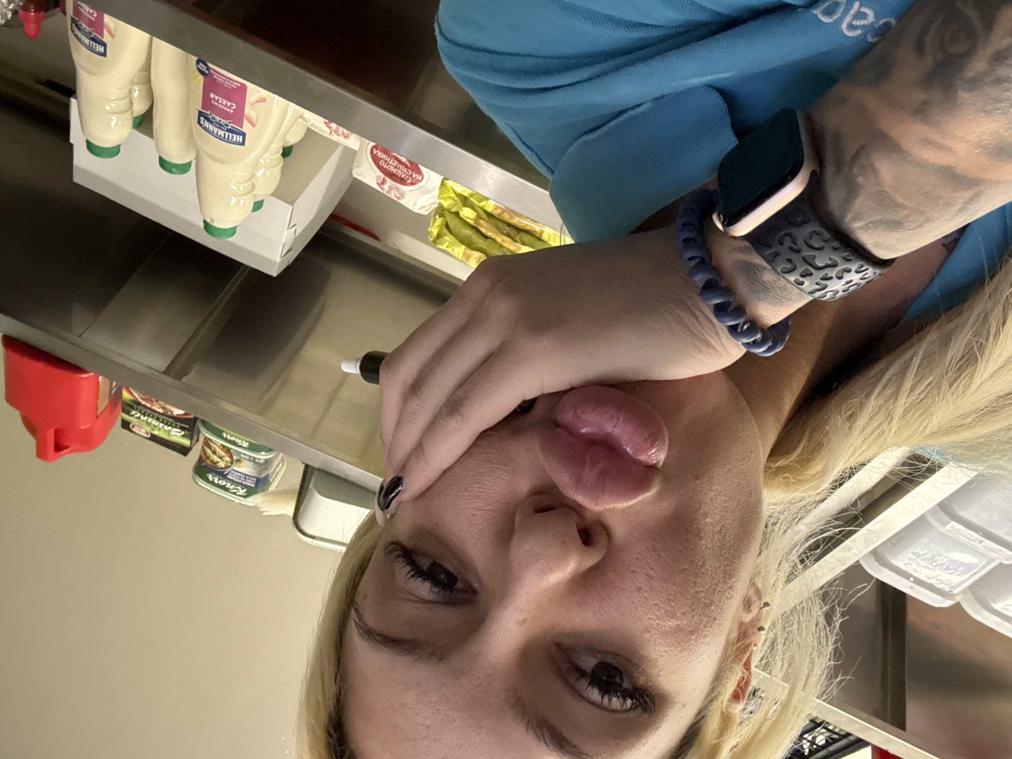 A close-up selfie shows a person with blonde hair and fair skin, with their head tilted and their tongue slightly out in a playful expression. One hand is raised, partially covering the side of their face, holding what appears to be a black marker or pen. The person has visible eyelash extensions or heavy mascara and dark nail polish on their index finger.
On the left arm, a smartwatch with a rose gold casing and a black patterned band is visible, along with a blue coiled wristband. Tattoos can be seen on the arm beneath the watch. The background suggests an indoor commercial setting, likely a food preparation area, kitchen, or storage room, in Plovdiv, Bulgaria.
Behind the person, various food items are arranged on shelves and counters. Bottles of "HELLMANN'S CAESAR" dressing are prominent, with green caps. Further back, white sachets labeled "ТРАЙНОТО НА САЛАТНИКА" (TRAINOTO NA SALATNIKA, likely meaning "The Long-lasting Salad" or similar, hinting at a salad component) are visible. Next to these are yellow-packaged items, possibly crackers or breadsticks. Other products include red containers and items branded "Knorr" in cans or packages. White plastic storage containers are also seen on the right side of the frame. The surfaces are primarily metallic, suggesting a professional kitchen environment. The overall mood conveyed by the subject is casual and lighthearted.
