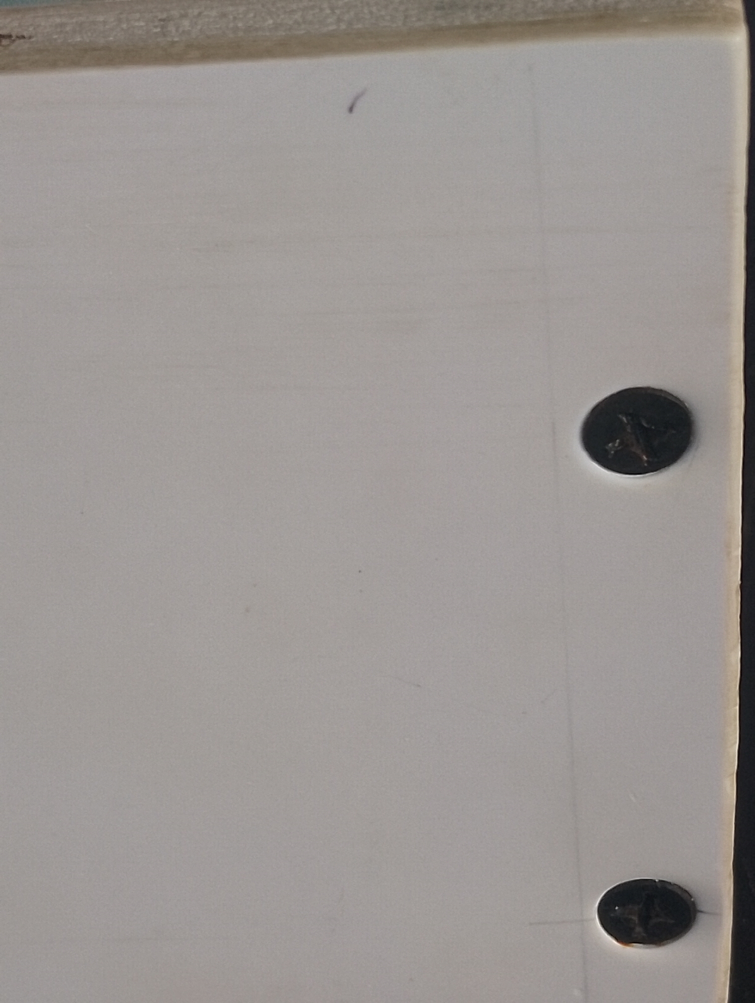 The image shows a close-up of a white surface with a slight texture, possibly a piece of furniture or a wall. Two dark-colored screws are visible, each with a Phillips head, embedded in the surface. The top screw is located towards the right edge of the image, and the bottom screw is positioned below it. There is a faint vertical line drawn on the surface between the two screws. A small, thin, curved purple mark is present in the upper left quadrant of the image. The top edge of the surface shows a lighter colored material, possibly wood, with a rough texture. The right edge of the surface is dark and appears to be a contrasting material. The image is likely a detail shot of an object or surface in an indoor setting, with no context of its location within a city.