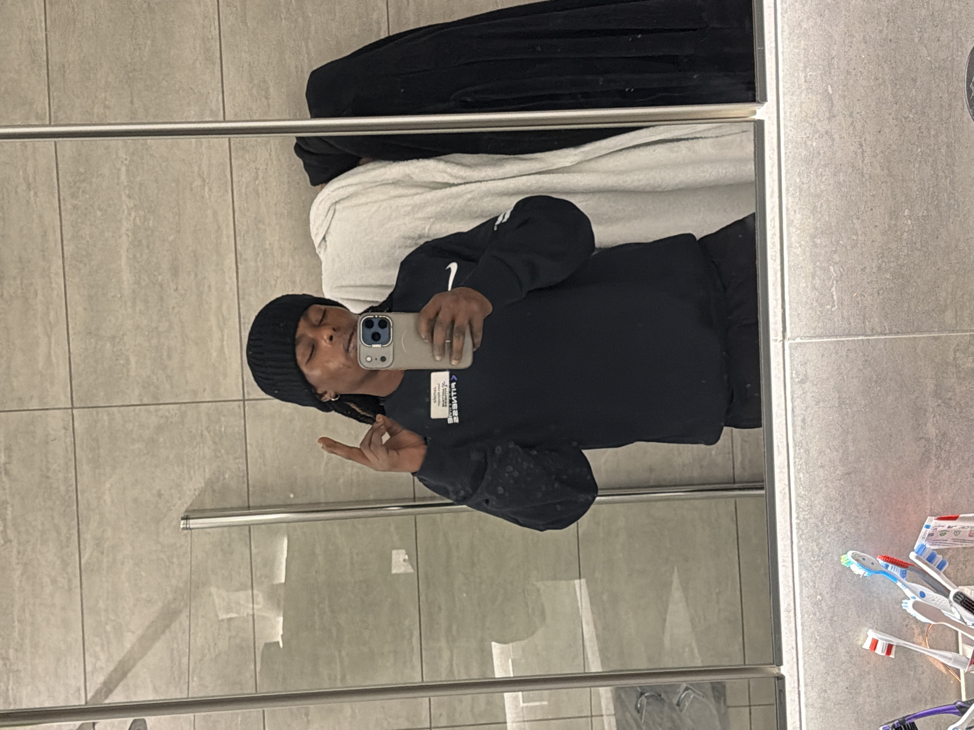 A person with dark skin and wearing a black beanie and black sweatshirt is taking a selfie in a bathroom mirror. They are holding an iPhone and making a hand gesture with their left hand. There is a white and black towel draped on a surface behind them. The bathroom has light grey tile walls and a glass shower door. There are toothbrushes mounted on the wall to the right. The location of the image is not discernible.