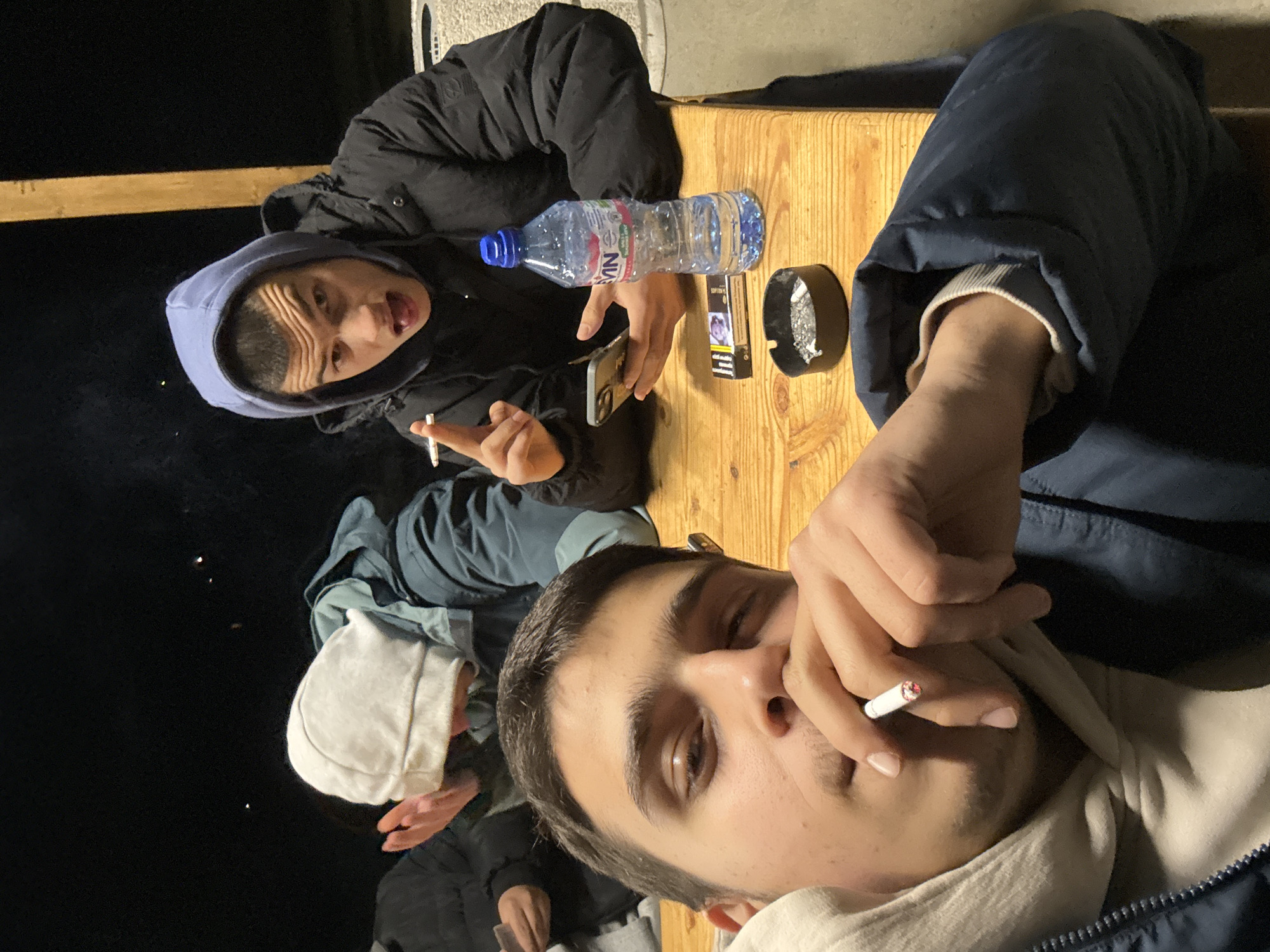 At night in Unknown, Unknown, the image shows a group of people gathered around a wooden table. One person is holding a lit cigarette to their mouth, while another person is also holding a lit cigarette and looking at the camera. On the table is a bottle of water, a pack of cigarettes, an ashtray, and a mobile phone. The environment is dark, with some light reflecting off the table and the subjects' faces.