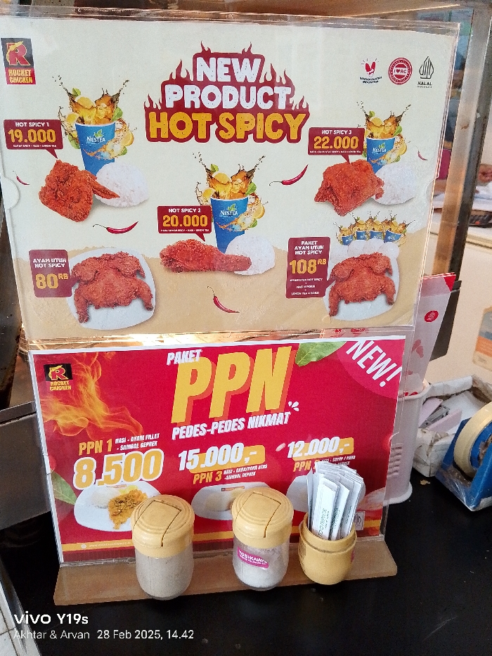 The image shows a food stall sign advertising "Hot Spicy" fried chicken. The sign features four different fried chicken dishes with different prices. The price is listed in Indonesian Rupiah. There is a large red sign at the bottom of the image promoting a "Paket PPN" meal option. The sign lists different packages and prices for the "Pedes-Pedes Nikmat" meal. The "Paket PPN" meal options include "PPN 1," "PPN 2," and "PPN 3," with prices of 8,500, 15,000, and 12,000 Rupiah, respectively. There are three small round containers in the foreground. The containers have yellow lids and appear to be filled with different condiments. The condiments include small white toothpicks and white powder. The sign is positioned on a black surface and partially obscures a blue and white patterned container in the background. The location of the image is in Patrol, Indonesia, at the coordinates -6.318 latitude and 107.986 longitude.