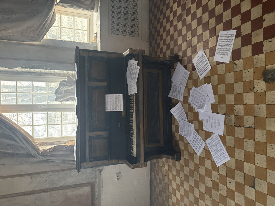 The scene is an interior room within an aged building, consistent with historical architecture found in Moscow, Russia. Centered in the mid-ground is a dark, upright piano exhibiting signs of wear, including discolored keys. A single sheet of musical notation is positioned on the piano's music stand, while additional sheets lie on the instrument's key cover. Approximately two dozen sheets of musical notation are scattered across the tiled floor, which features a checkerboard pattern of reddish-brown and beige squares showing extensive wear and dirt.
Three large, multi-paned windows dominate the background, admitting significant natural light and revealing blurred outdoor foliage and architectural elements. Faded, patterned curtains flank the windows. The room's walls are light-toned with extensive peeling paint and plaster exposing underlying material. A white double electrical outlet is affixed to the left wall. An ornate, light-colored metal radiator is located beneath the right window. Two light-colored sculptural elements, one resembling a draped form and another an abstract shape, are situated on the windowsill between the left and central windows. No persons are depicted.