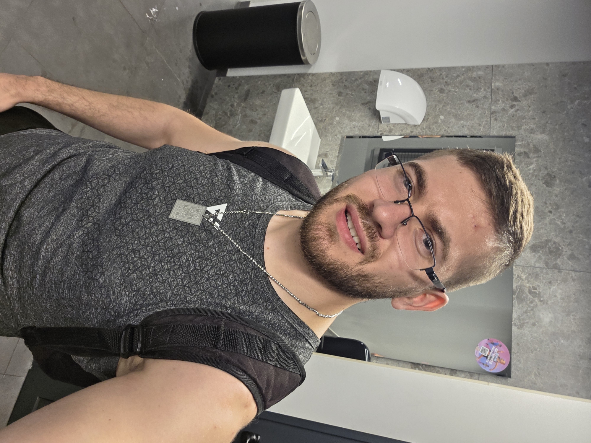 A man with a beard and glasses is taking a selfie in what appears to be a restroom. He is wearing a gray patterned tank top and a black backpack. A silver chain with a pendant hangs around his neck. In the background, there is a black trash can, a white sink and hand dryer, a mirror with a sticker on it, and gray tiled walls. The location is not discernible from the image.