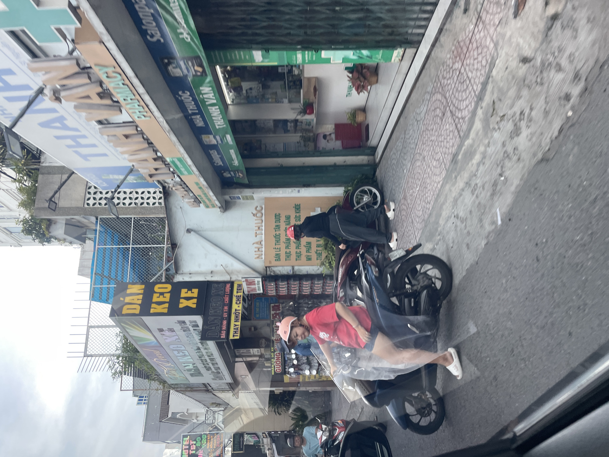 The image depicts a street scene in Thủ Đức, Vietnam, likely in the morning or late afternoon due to the lighting. A woman wearing a red t-shirt, blue shorts, and a pink helmet with rabbit ears is seated on a dark-colored motorcycle. She is holding the handlebars and looking forward. To her left, another person on a motorcycle is partially visible. In the background, commercial establishments line the street. One prominent sign reads "DÁN KEO XE" which translates to "Motorcycle Sticker Application." Another sign for "THUOC TAY" (medicine) with the name "THANH VAN" is visible, suggesting a pharmacy. The buildings are multi-story structures with visible signage and awnings. The street itself is paved, with a tiled sidewalk running alongside the buildings. There are other motorcycles and possibly some debris on the sidewalk. The overall atmosphere is that of a busy urban commercial area.
