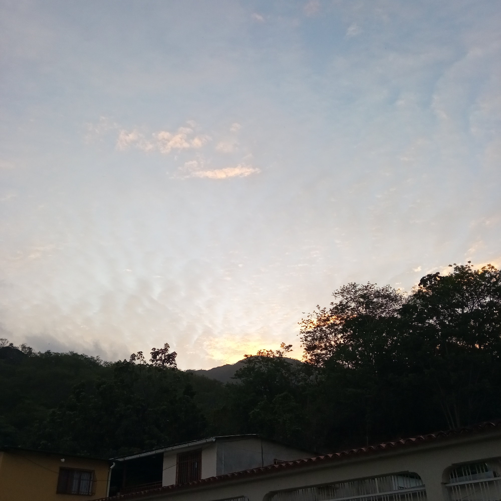 The photograph captures a scene in El Limón, Venezuela, depicting the sky at sunset above a landscape with a mountain range and foliage in the middle ground. The foreground shows rooftops of buildings, including a house with a window and a tiled roof. The sky has a gradient of colors, with the sun setting behind the mountains, creating a bright glow. The cloud cover is diverse with some wispy clouds in the lighter areas.