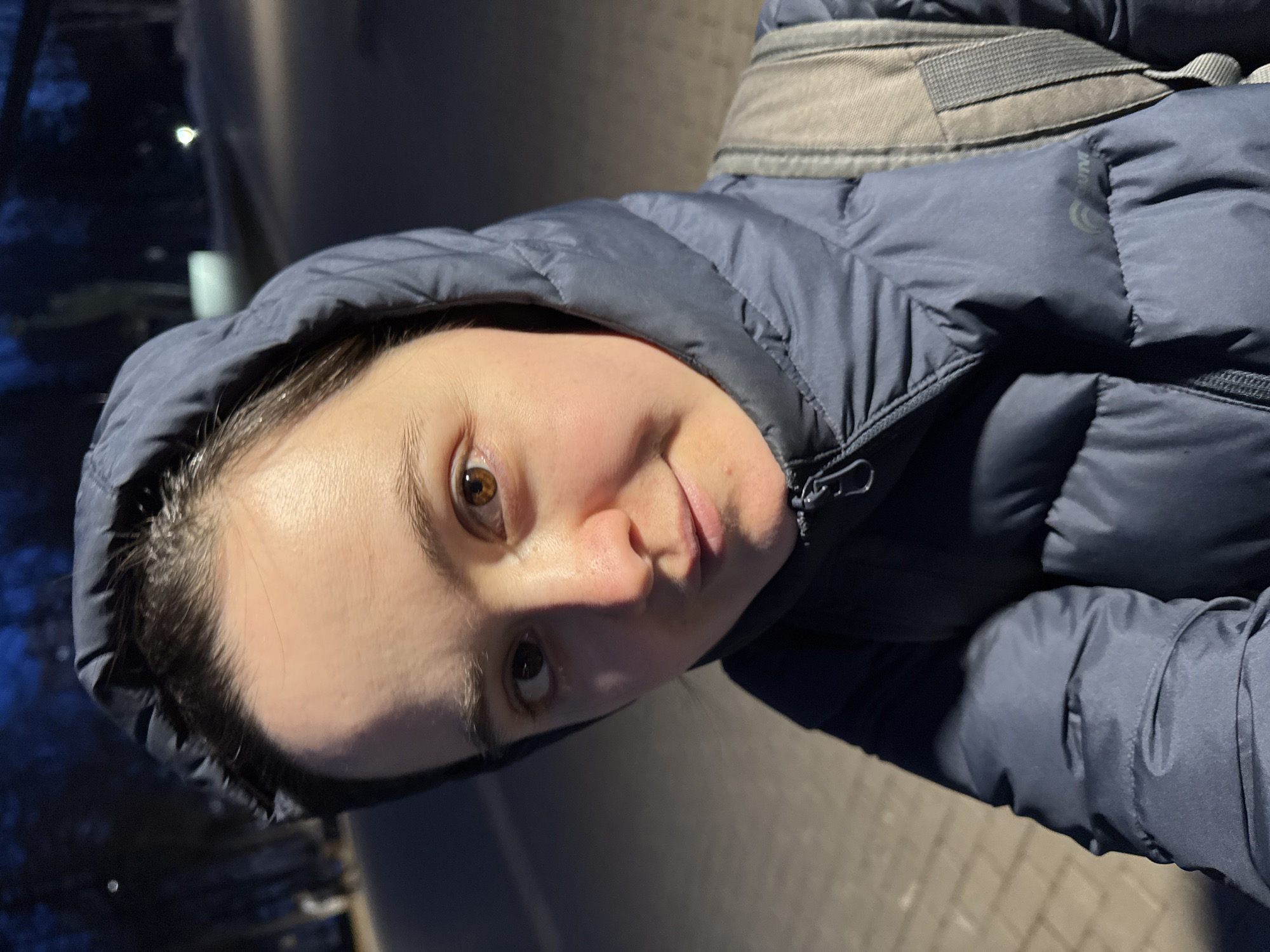 A close-up, tilted selfie shows a woman wearing a dark blue hooded puffer jacket. The hood is up, framing her face. She has light skin, brown eyes, and dark hair. A gray backpack strap is visible over her shoulder. The background appears to be an urban environment at night, with blurry hints of streetlights and trees. Given the location in Izhevsk, Russia, it is probable that the pavement is the sidewalk.