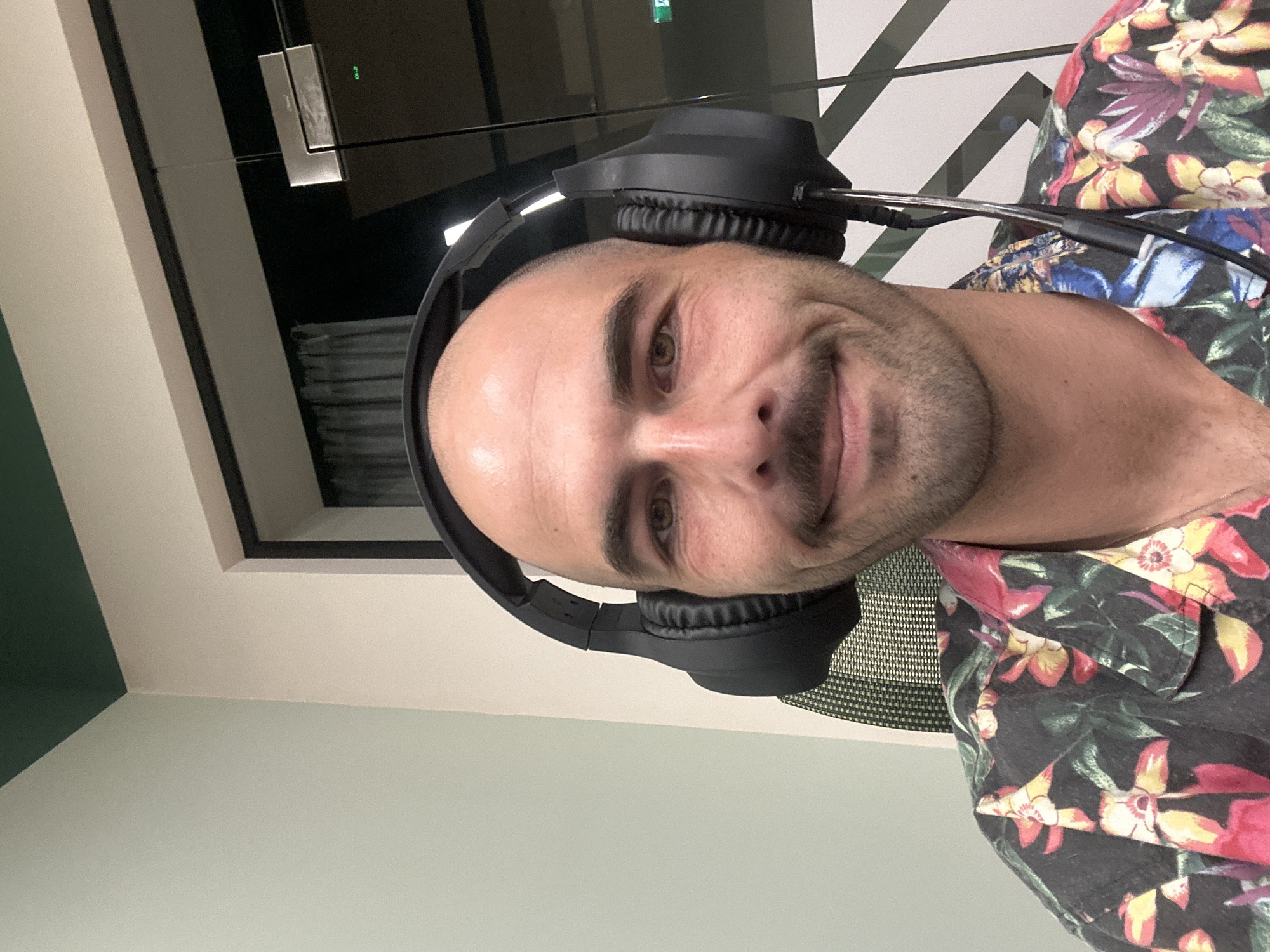 A close-up, slightly angled portrait shows a light-skinned, bald man wearing black headphones and a colorful floral shirt. He is smiling slightly and looking directly at the camera. The background consists of a neutral-toned wall and a window. No specific information indicates it is Budva, Montenegro.
