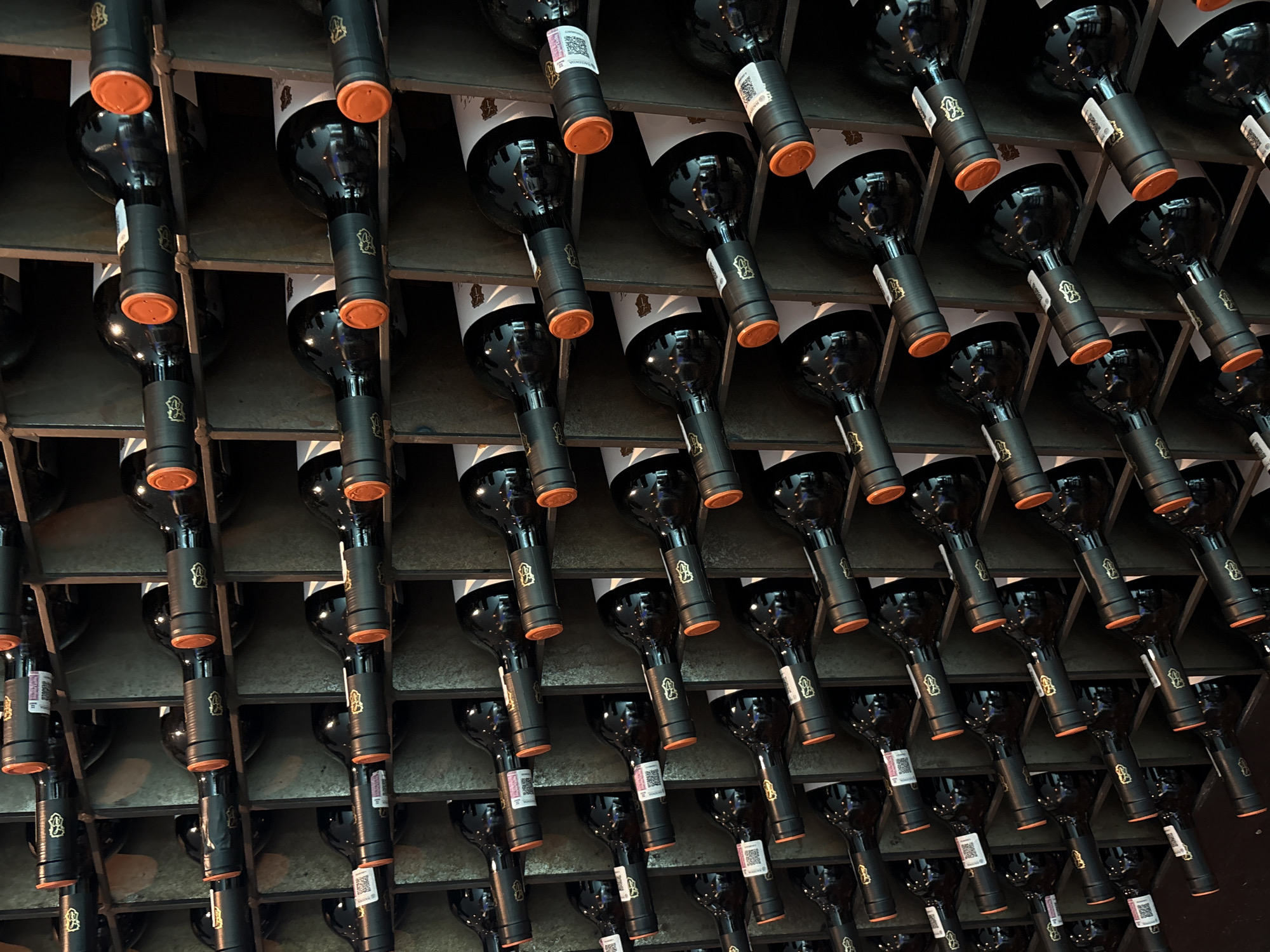 The image shows a close-up, upward-angled view of a wine rack filled with numerous dark-colored wine bottles. The bottles are positioned horizontally within a metal grid system. The bottom parts of the bottles are visible, with dark capsules and labels. Many of the capsules are orange, while the labels on some bottles are covered with bar codes. The location is Huixquilucan de Degollado, Mexico.