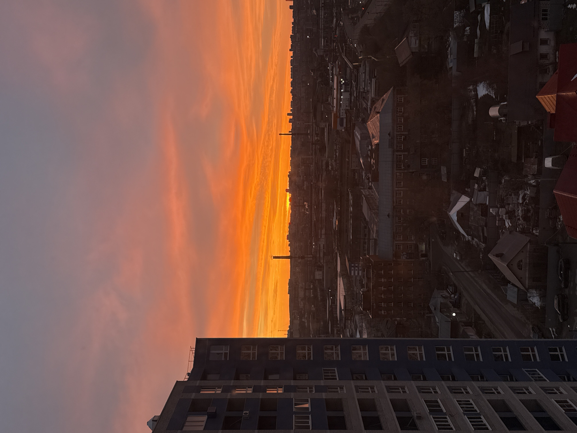 The image shows a sunset view over the city of Izhevsk, Russia. In the foreground, the corner of a multi-story building is visible, featuring a grid-like facade of windows. The sky transitions from a muted orange near the horizon to a soft gray-pink towards the top. The sun casts a vivid orange light through a narrow band in the middle distance, illuminating the cityscape's structures, including residential buildings, industrial facilities with a tall smokestack, and a network of roads. The light fades as one looks towards the horizon from left to right, with buildings growing darker. There are no people visible in the image.