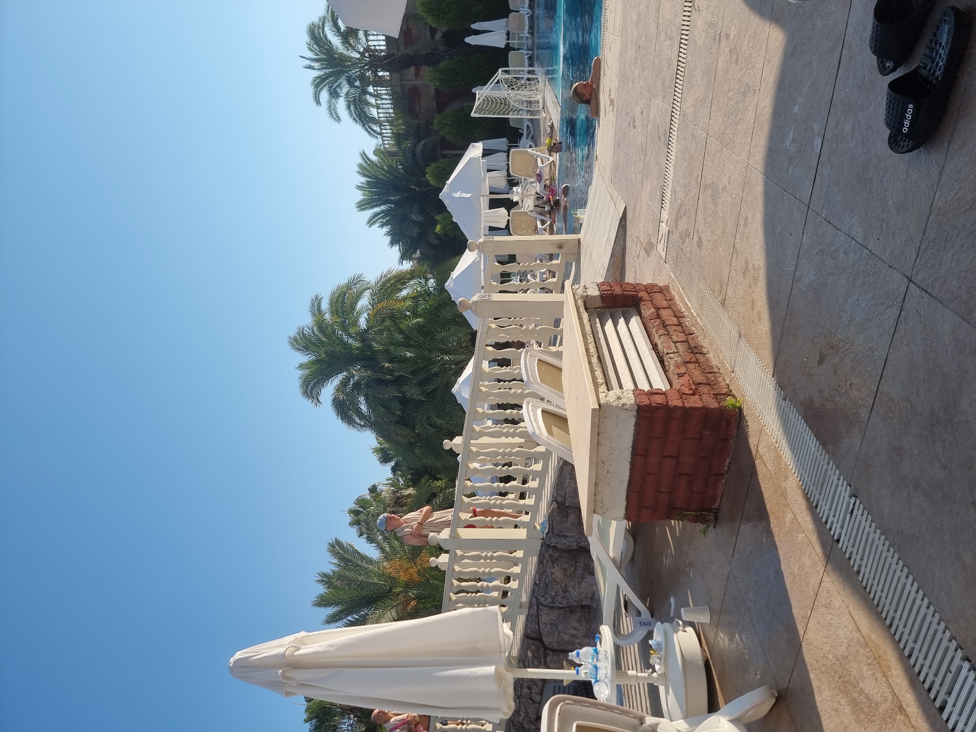 The image captures a sunny day at a resort pool in Manavgat, Turkey. The foreground is dominated by a tiled patio with a prominent white drain grate. A white lounge chair and a small white table with bottles of water are visible on the left. In the middle ground, a brick and stone structure with a white slatted bench sits adjacent to the pool. A white railing with ornate balusters runs along a raised walkway. Palm trees and other lush greenery form the backdrop.
Several people are present. On the walkway, a person wearing a towel around their neck and shorts stands near the railing. Further back, a few individuals are relaxing in lounge chairs under white umbrellas. In the pool, a woman is partially submerged, leaning on the edge, and another person can be seen in the water. A soccer goal net is positioned at the edge of the pool. On the far right, a pair of black Adidas slides lie on the tiled ground. The sky is clear blue, indicating good weather. The overall scene suggests a leisurely vacation setting.