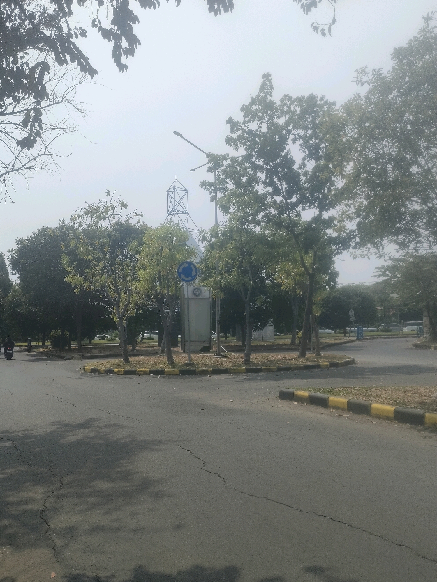 The image depicts an outdoor scene at a roundabout in Surabaya, Indonesia, likely a campus or park area. In the foreground, a paved road shows signs of cracking and is partially covered by the dappled shadows of trees. A motorcycle rider is visible on the left side of the frame, moving away from the camera.
The roundabout itself is bordered by a curb painted with alternating yellow and black stripes. Within the roundabout, several trees of varying heights and foliage densities are present. A tall metal structure, possibly a tower or part of an antenna, is visible in the background, partially obscured by trees and hazy sky. A street light is also present, mounted on a pole. In the middle of the roundabout, a sign with a circular blue background and a white arrow indicating a roundabout is mounted on a pole next to a rectangular grey utility box.
The environment appears somewhat hazy, suggesting a warm or humid climate, typical of Surabaya. Trees line the surrounding area, with some cars visible in the background, indicating a road network beyond the immediate scene. The overall impression is of a public or institutional space with maintained landscaping.