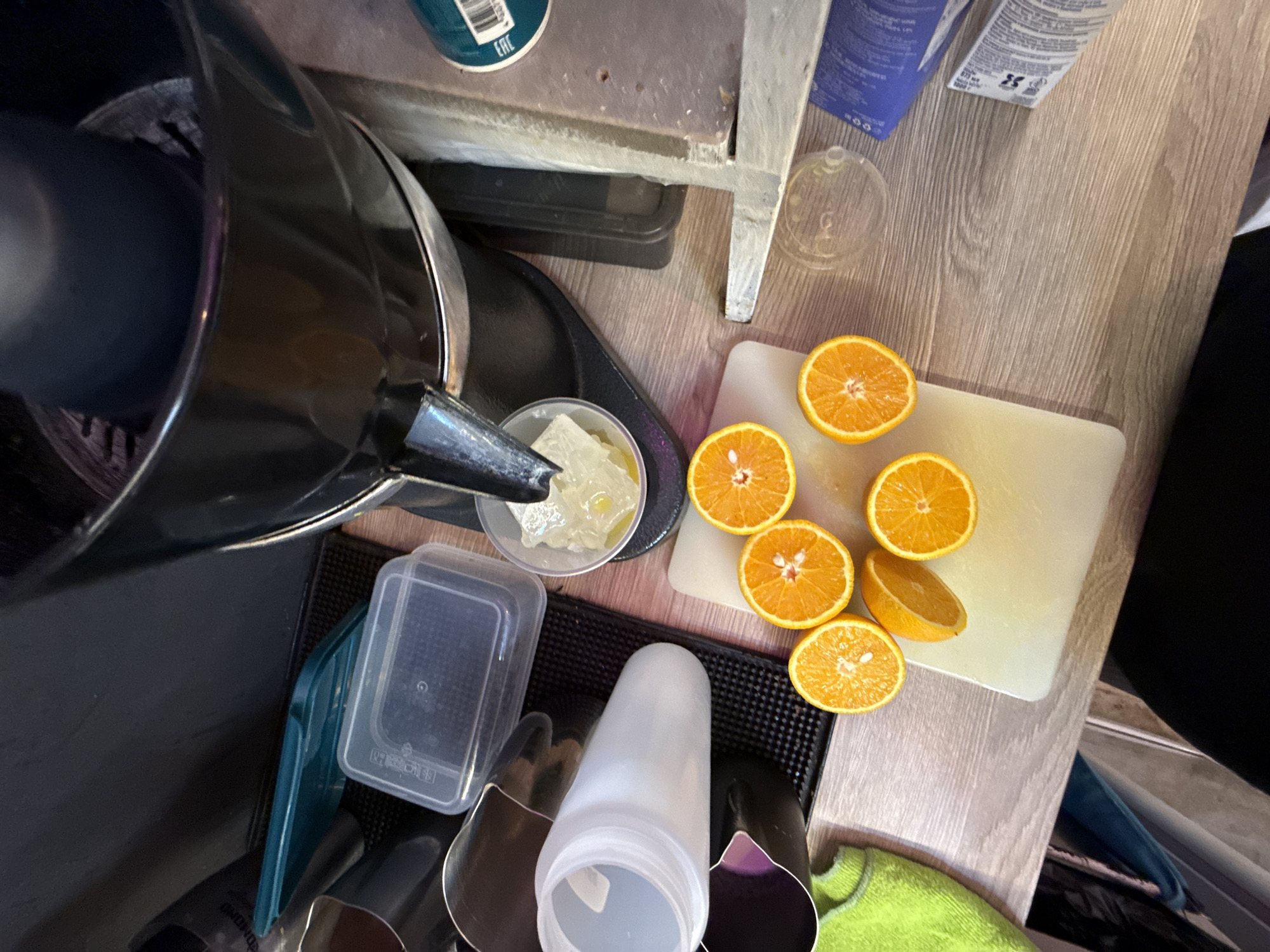 The image depicts a close-up, overhead view of a kitchen counter. In the center, a black electric citrus juicer is positioned with its spout pouring a pale yellow liquid into a small white bowl containing ice cubes. Surrounding the juicer, several sliced oranges are arranged on a white cutting board. To the left of the juicer, a translucent white bottle and a turquoise plastic container are visible. The counter surface is a light-colored wood grain. To the right of the juicer, part of a stainless steel appliance and various cleaning supplies are present. The overall scene suggests the preparation of fresh juice. The image is taken from a high angle, looking down. There is no indication of the location being in Moscow, Russia.