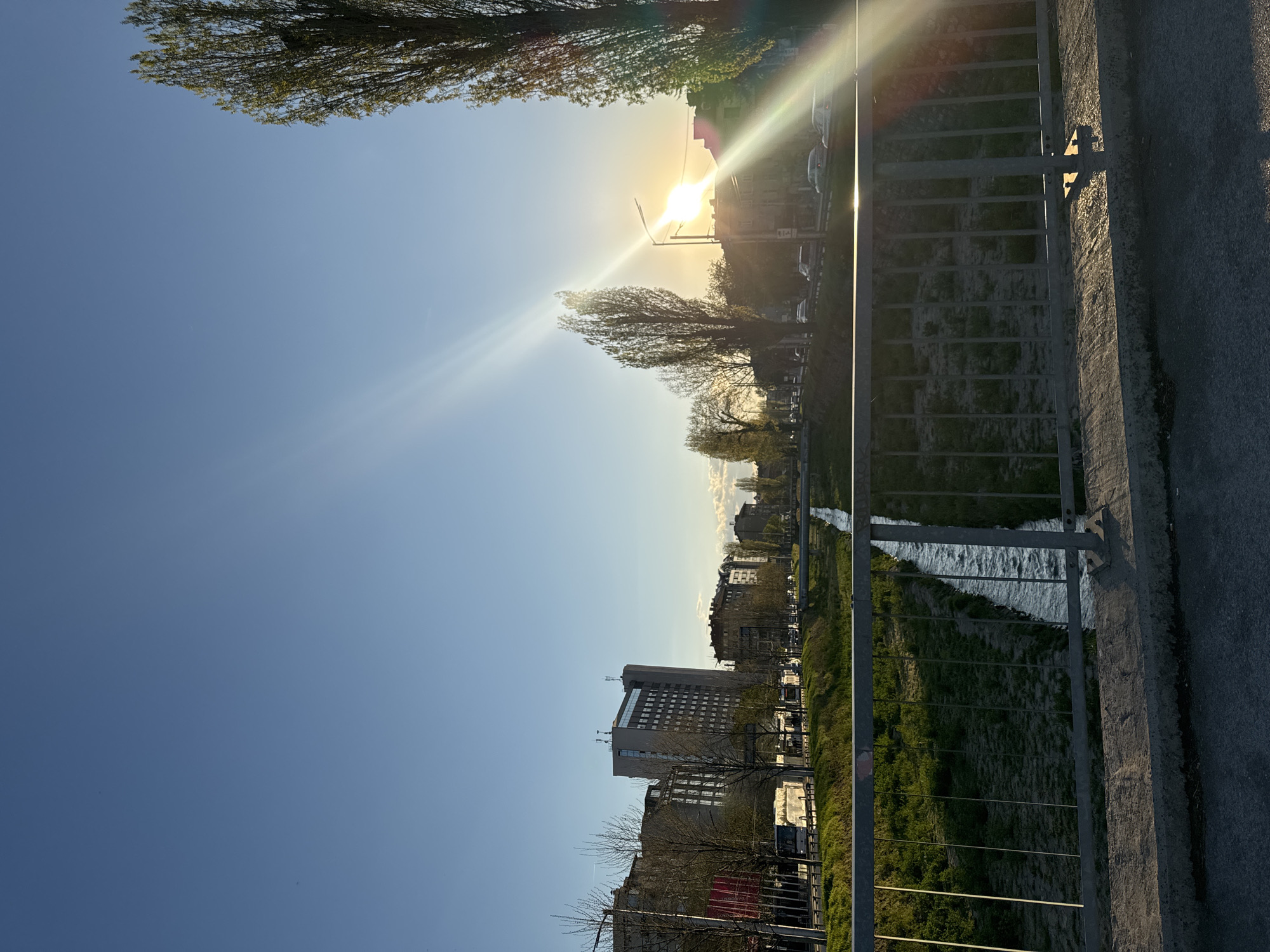 The image depicts a landscape in Sofia, Bulgaria at sunset. The view is from a bridge overlooking a waterway. A metal railing runs along the right edge of the frame. Beyond the railing is a grassy bank, and a canal with flowing water. Buildings line the opposite side of the canal, including a tall, multi-story office building. The sun is setting behind the buildings and trees, creating a bright glare and lens flares. The sky is blue. Trees are visible along both sides of the waterway. Cars and other vehicles are parked along the street adjacent to the buildings.