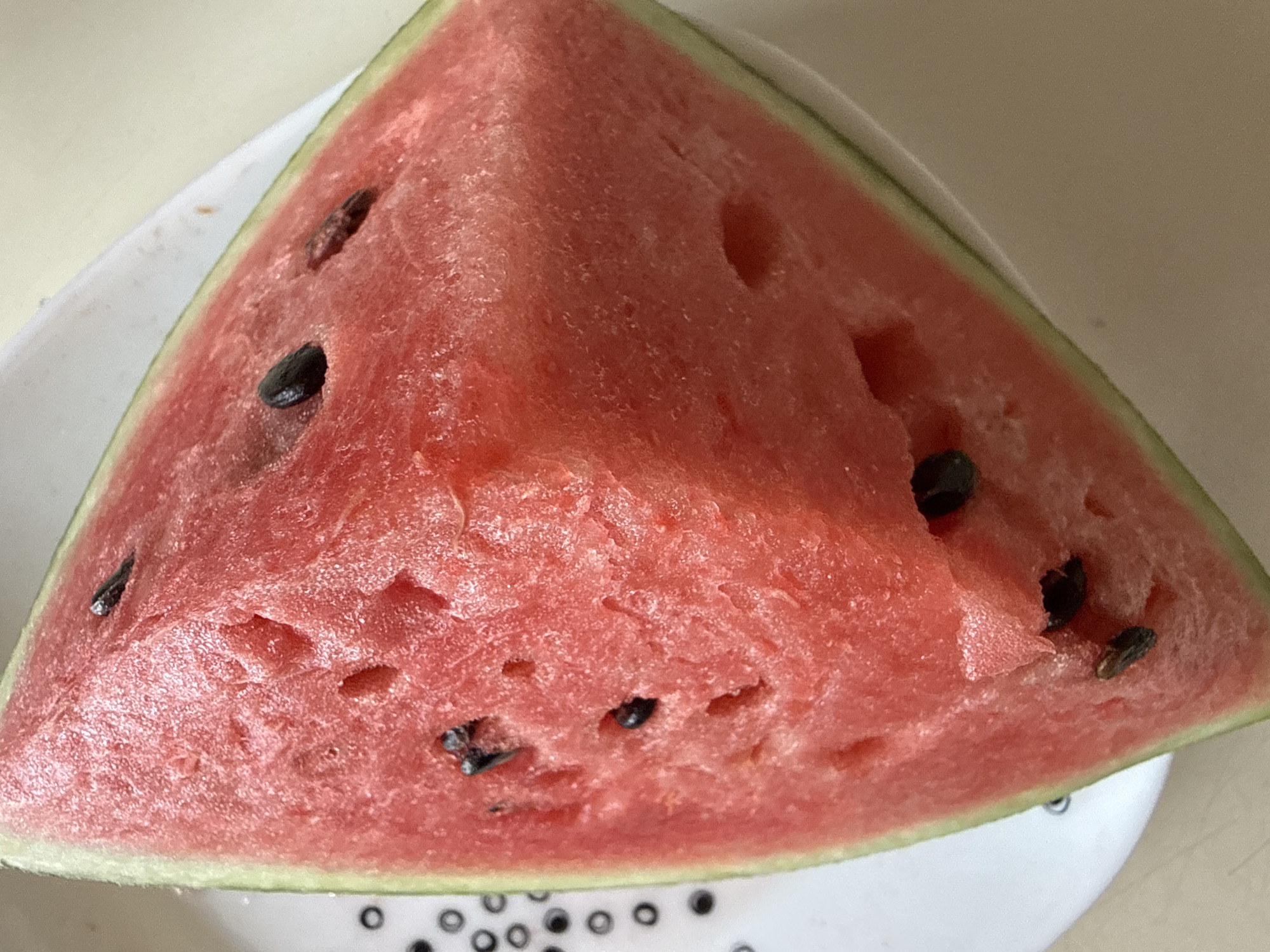 A close-up, overhead shot shows a triangular slice of watermelon resting on a white plate with a decorative pattern. The watermelon flesh is a vibrant red, with a slightly textured surface and visible juice. Several black watermelon seeds are embedded within the flesh. The rind is visible along the outer edge, a lighter green color. The plate appears to be ceramic and sits on a light-colored surface, possibly a table. The background is blurred, with hints of the environment. The image does not contain any information related to the city of Okhtyrka, Ukraine.