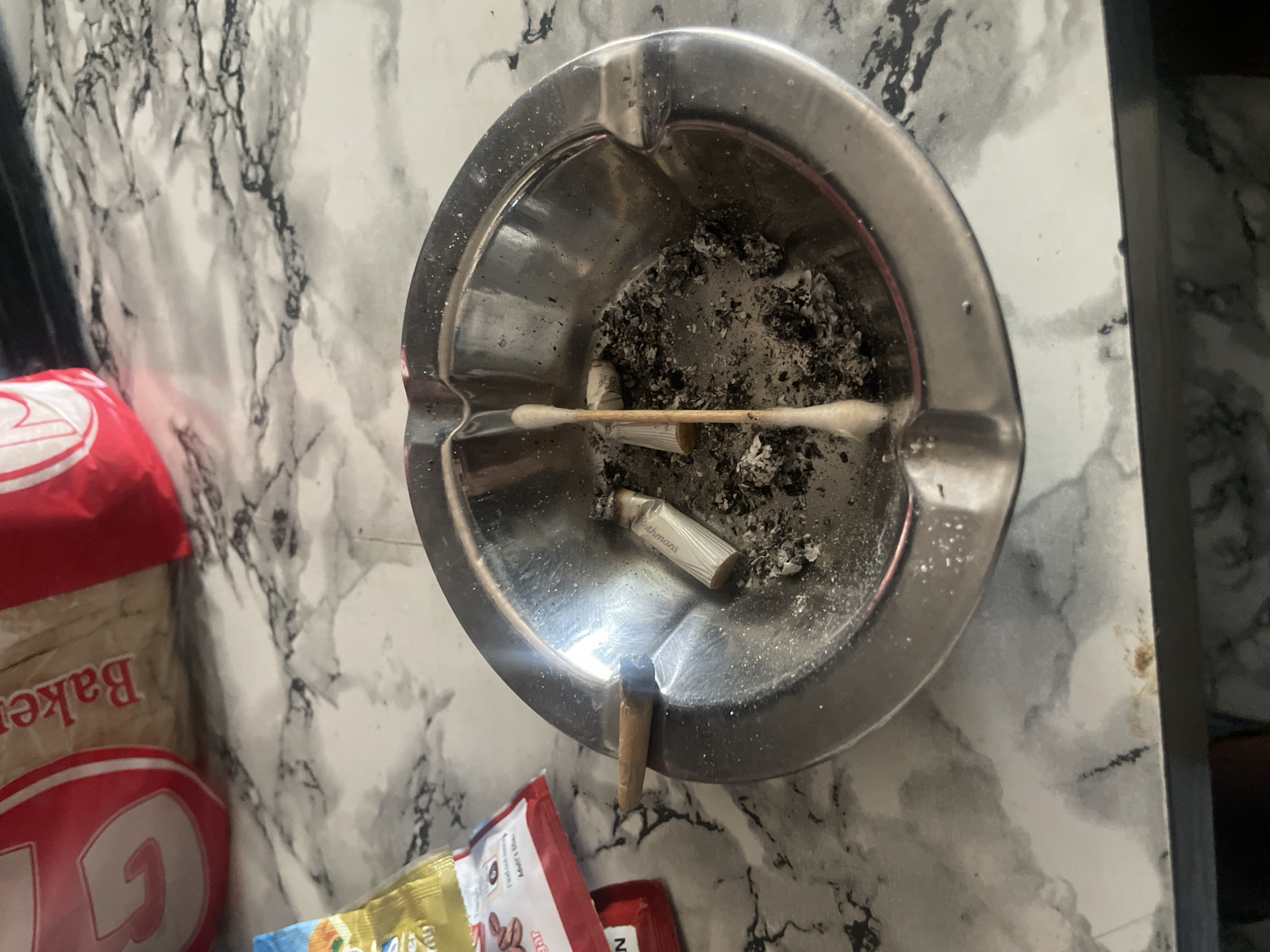 The image depicts an ashtray filled with cigarette butts and ashes, situated on a marble-patterned countertop. A cotton swab is placed vertically across the ashtray, with one end resting on a cigarette butt. To the left of the ashtray, a portion of a bag of snacks is visible. The background shows a red and white bag, likely containing baked goods, with the word "Bakery" printed on it. The lighting suggests an indoor setting. The image does not contain any information about the city of New Stadium, Nigeria.