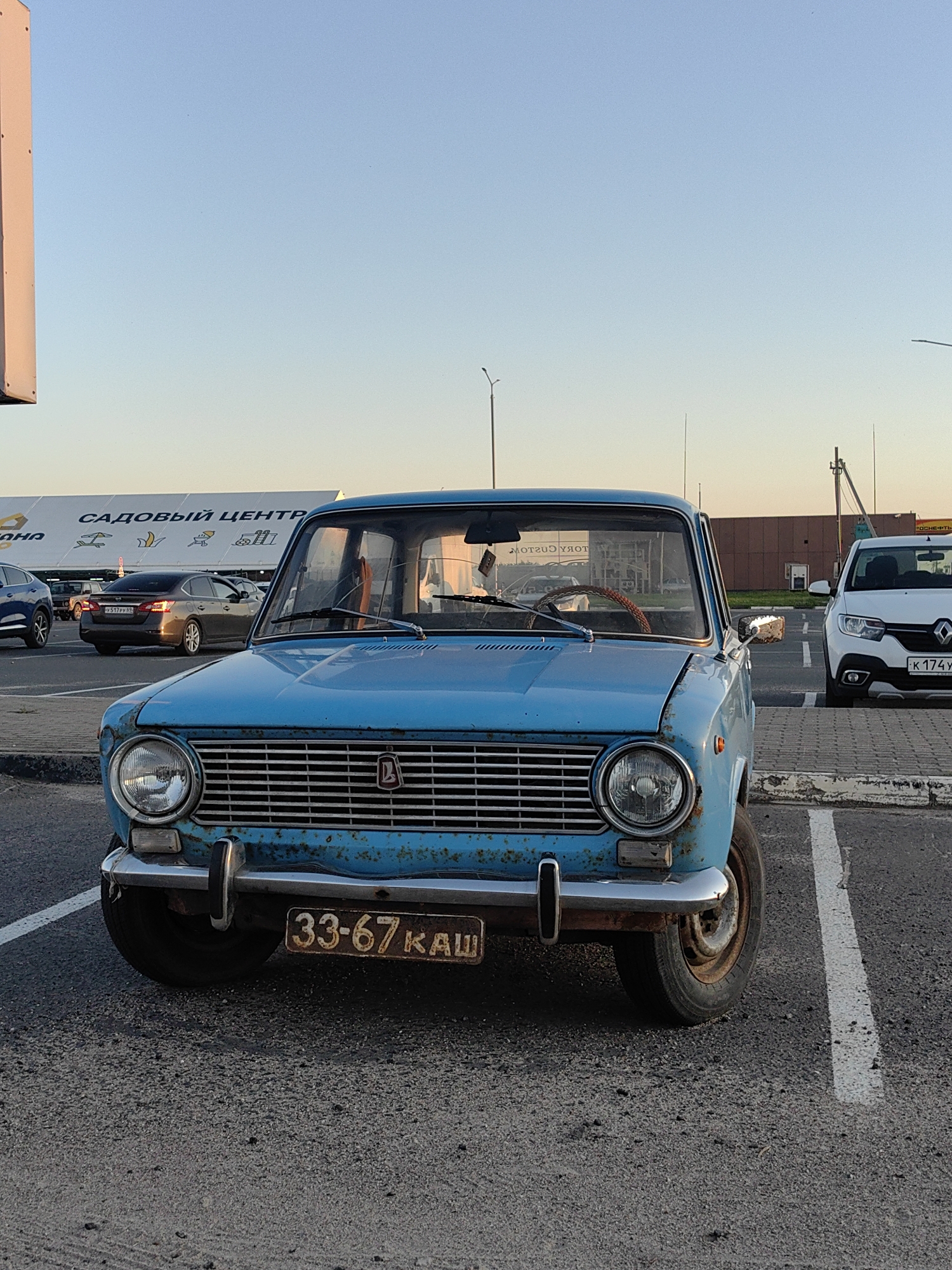 A faded blue vintage Soviet-era car, likely a Lada, is parked in a lot, facing the camera. The car exhibits significant rust and wear. A license plate reading "33-67 КАШ" is affixed to the front bumper. In the background, a large building with the text "САДОВЫЙ ЦЕНТР" (Garden Center) is visible, suggesting a retail or commercial area. Other vehicles are present in the parking lot, including a white modern car to the right and a darker sedan further back. The scene is set during daylight, with a clear sky, indicative of a temperate climate. The location is identified as Tver, Russia, a city situated on the Volga River.