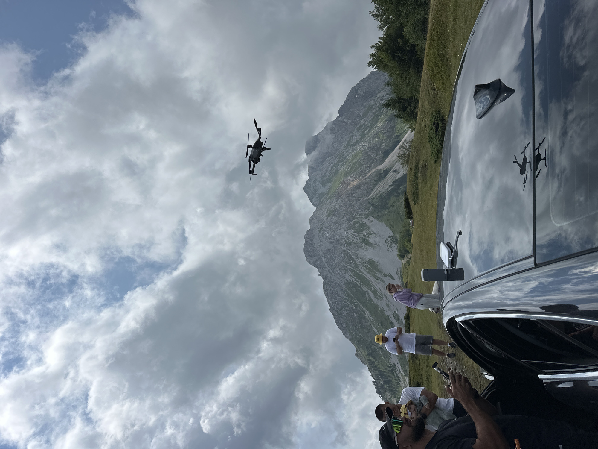 In the mountain landscape of Krajišta, Montenegro, a group of five people stands near a black car. In the foreground, a man in a black t-shirt and sunglasses holds a drone controller. To his left, another man, partially obscured, stands with a baby. Behind them, a man in a yellow hat and shorts looks at his phone, and a woman in a purple hoodie also gazes down. A white dog stands near the woman. A drone is visible in the sky above the rugged mountain peaks. The sky is filled with dramatic, cloudy formations. The car's roof reflects the cloudy sky and the drone.