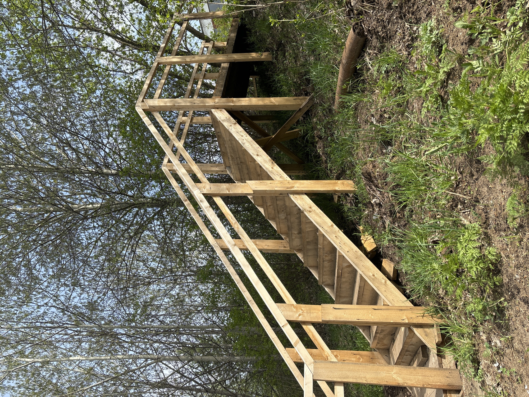 A wooden staircase ascends a grassy slope near Chaikovsky, Russia. The unfinished structure features steps, railings, and a landing platform at the top. Trees are visible in the background. The hillside has a mix of grass, weeds, and bare dirt.