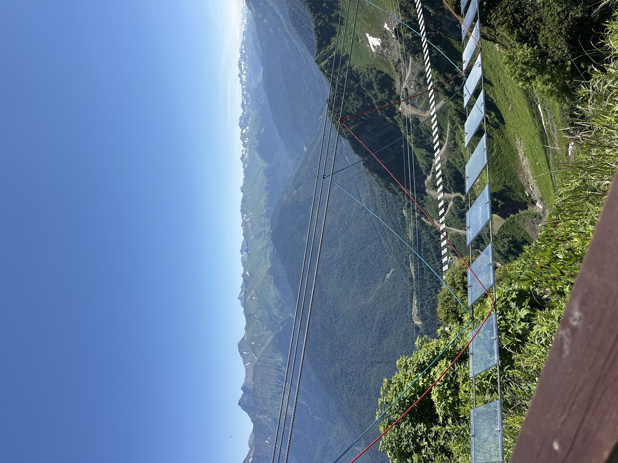 This is a photograph of a mountain landscape in Estosadok, Russia, on a clear, sunny day. The foreground features a wooden railing and lush green vegetation. A suspended bridge with clear glass panels spans across the frame, supported by thick cables and red and blue ropes. Beyond the bridge, a vast expanse of green, forested mountains stretches into the distance. In the background, snow-capped peaks rise against a clear blue sky. The scene suggests a popular tourist destination, likely an observation point or adventure park, offering panoramic views of the Caucasus Mountains.