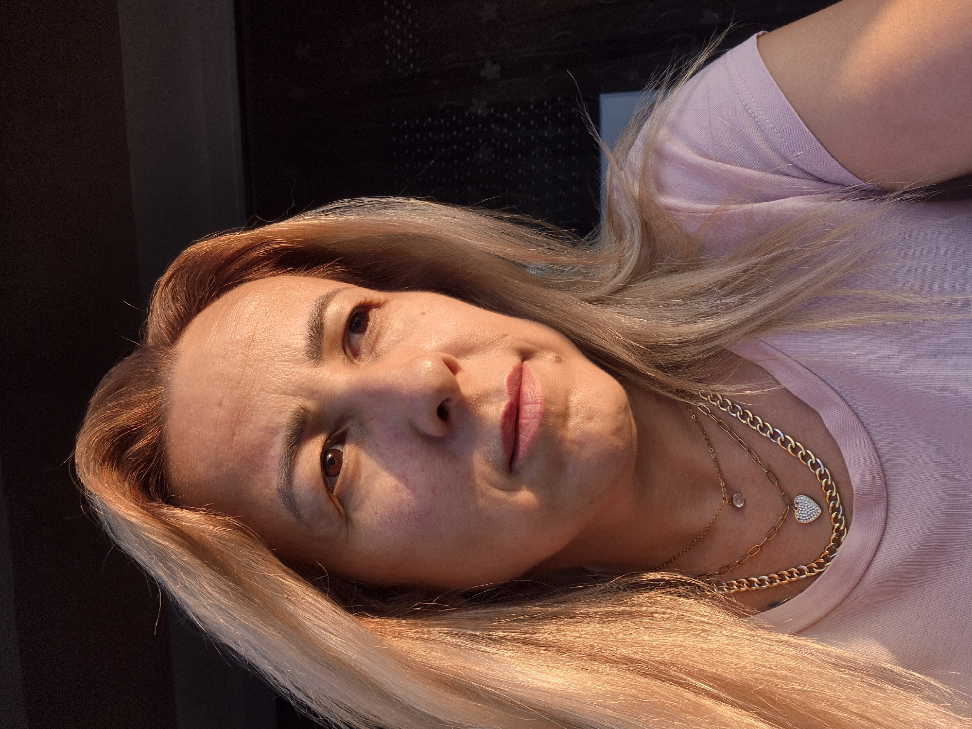 A head and shoulder shot of a woman with blonde hair, illuminated by the warm glow of what appears to be sunset light. She is looking off to the right, her expression thoughtful. She is wearing a pink t-shirt and multiple gold necklaces, one of which has a heart-shaped pendant covered in small white stones. The background is dark and out of focus, with some indistinct patterns visible.