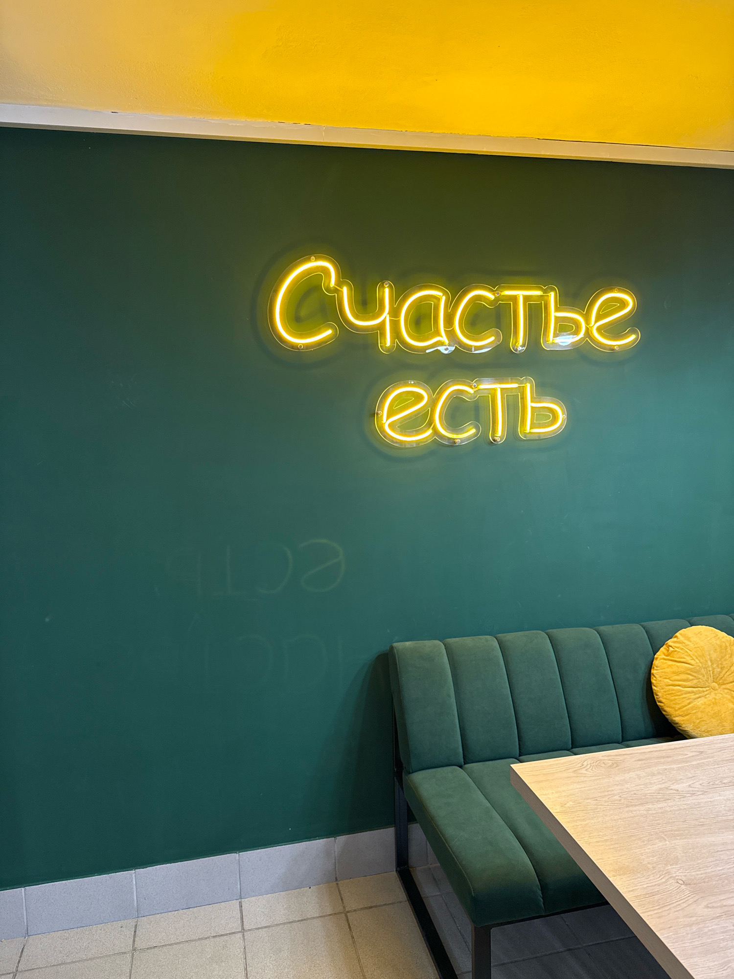 The image depicts an interior scene with a neon sign on a dark green wall. The neon sign, glowing yellow, reads "Счастье есть" which translates to "Happiness exists" in English. Below the sign, a dark green upholstered bench seat with a tufted design is visible, complemented by a yellow circular cushion. A light-colored wooden table partially extends into the frame from the right. The wall above the green section is painted yellow. The floor in the foreground is tiled with light beige squares. Faint, almost imperceptible, lighter text is visible on the green wall below the neon sign, possibly remnants of previous decor or ghosting. The lighting suggests an indoor setting, possibly a cafe or restaurant. There are no people present in the image. The location is stated to be in the city of Chaikovsky, Russia.