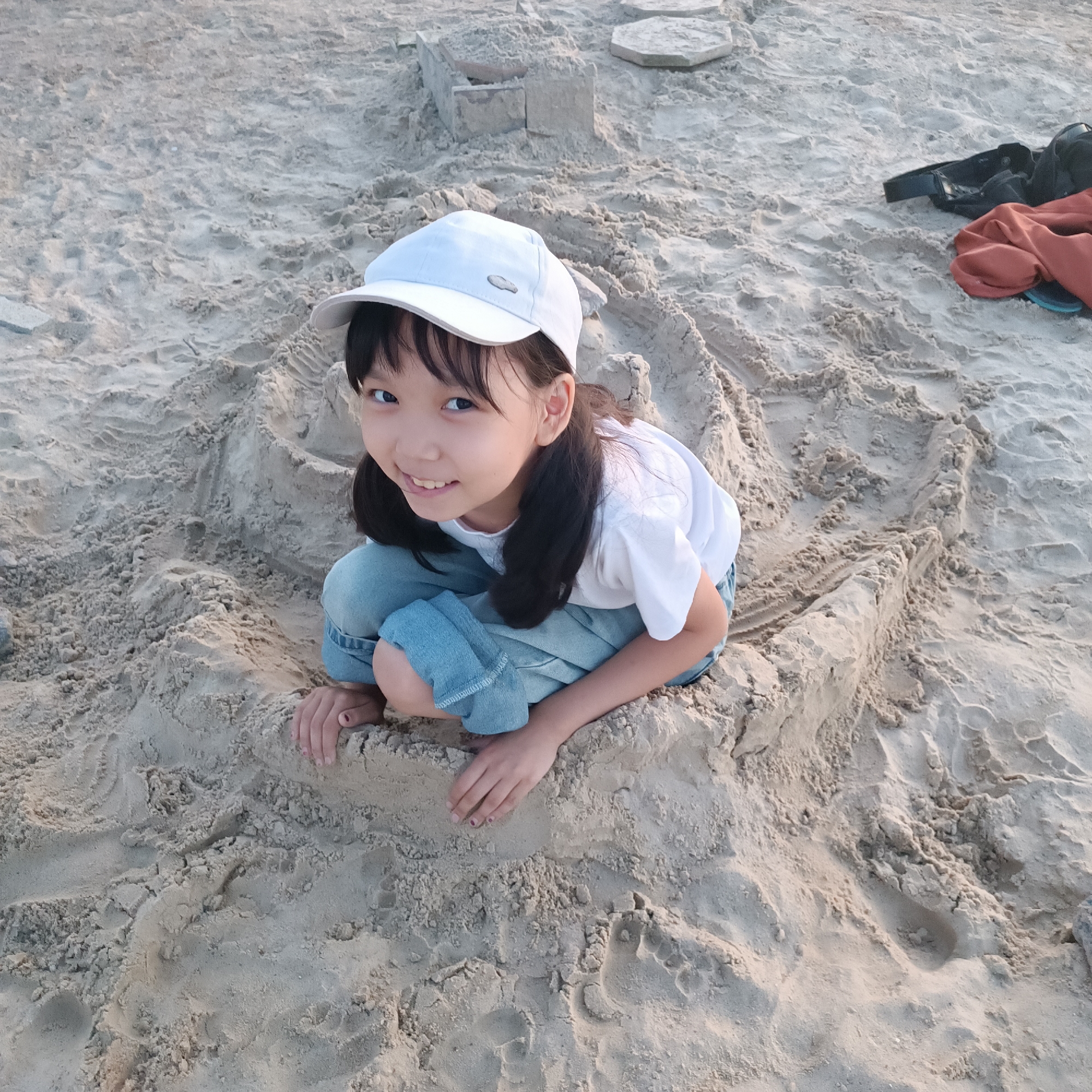 A young girl wearing a white baseball cap and blue jeans is crouching on a sandy beach, playing with sandcastles. She has dark hair pulled into pigtails and is smiling at the camera. The sand is light brown and appears wet in places, with footprints and the remnants of sand structures visible. In the background, there are more sand formations, and to the right, a black bag and an orange cloth are lying on the sand. The lighting suggests it is late afternoon or early evening, with a soft, diffused light. The scene evokes a sense of childhood fun and relaxation at the beach.