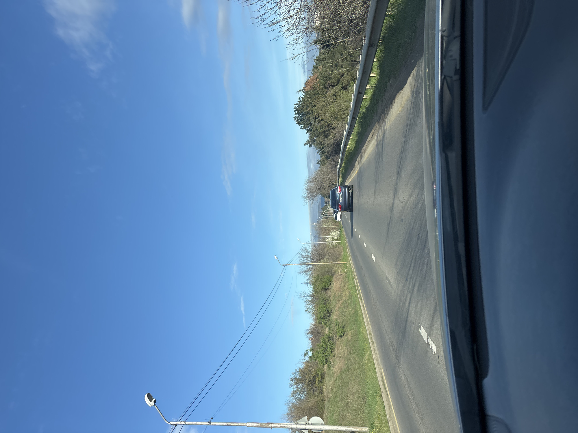 The scene depicts a view from inside a vehicle, likely a car, moving along a paved road. Another vehicle, a dark-colored car, is visible in the distance, ahead on the same road. The road is bordered by green vegetation on both sides, with trees and shrubs lining the route. Overhead, utility poles and power lines are noticeable against a clear blue sky with sparse clouds. The location appears to be outside of a densely populated urban area, with the surrounding environment suggesting a more suburban or rural setting. There are no visible signs indicating that it is in Tbilisi, Georgia.