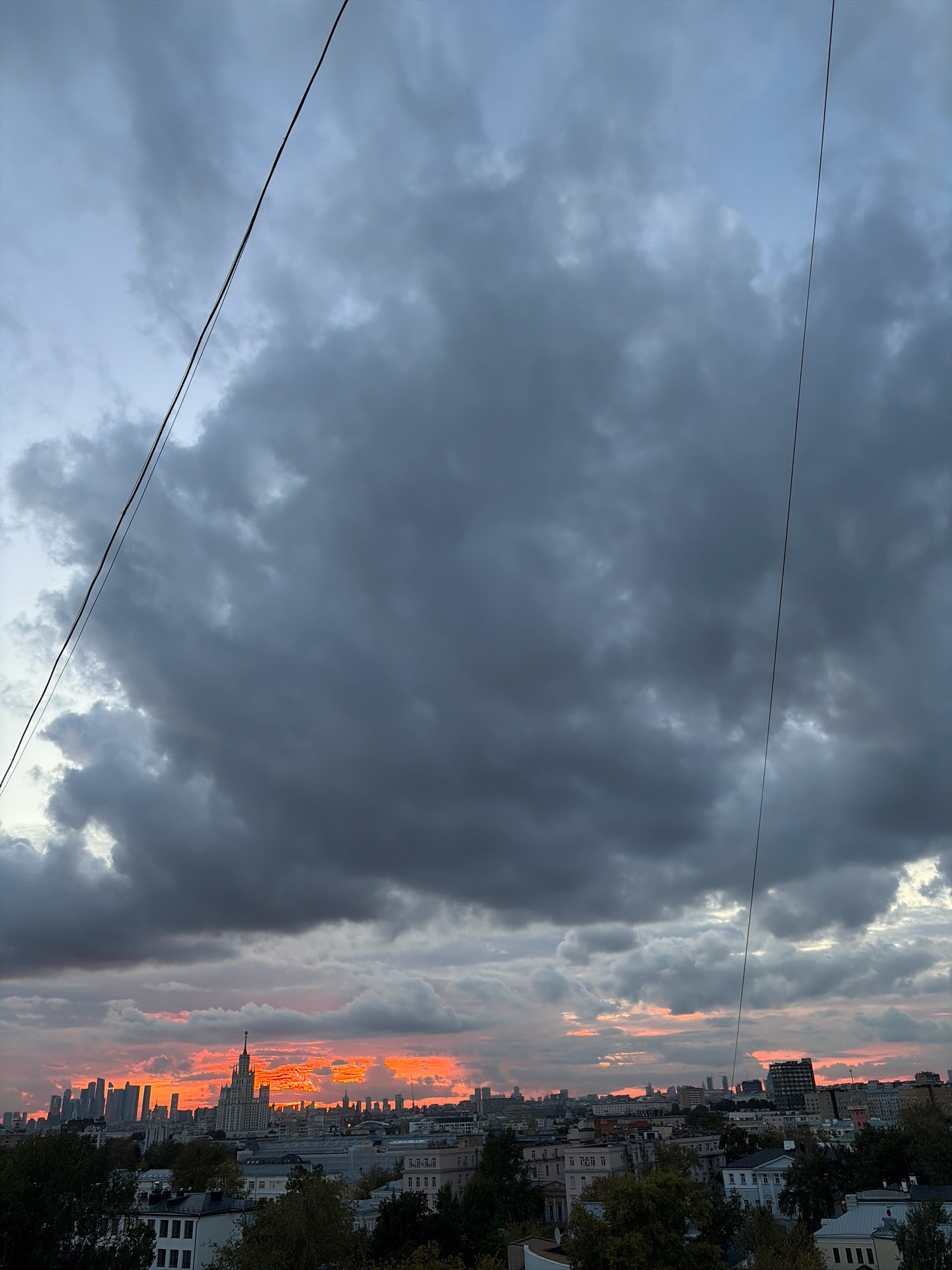 A wide shot captures a view of the Moscow skyline at sunset, with a dramatic sky filled with dark, heavy clouds. The sky is illuminated by a vibrant orange and pink glow peeking through the clouds, creating a stark contrast between light and dark. In the foreground, several buildings of varying heights and architectural styles are visible, interspersed with trees that are beginning to show autumn colors. A prominent Stalinist skyscraper, likely the Kudrinskaya Square Building, stands out among the cityscape. Two thin electrical wires run diagonally across the upper portion of the frame, obscuring parts of the sky. The overall impression is of a city preparing for evening under a stormy yet colorful sky.