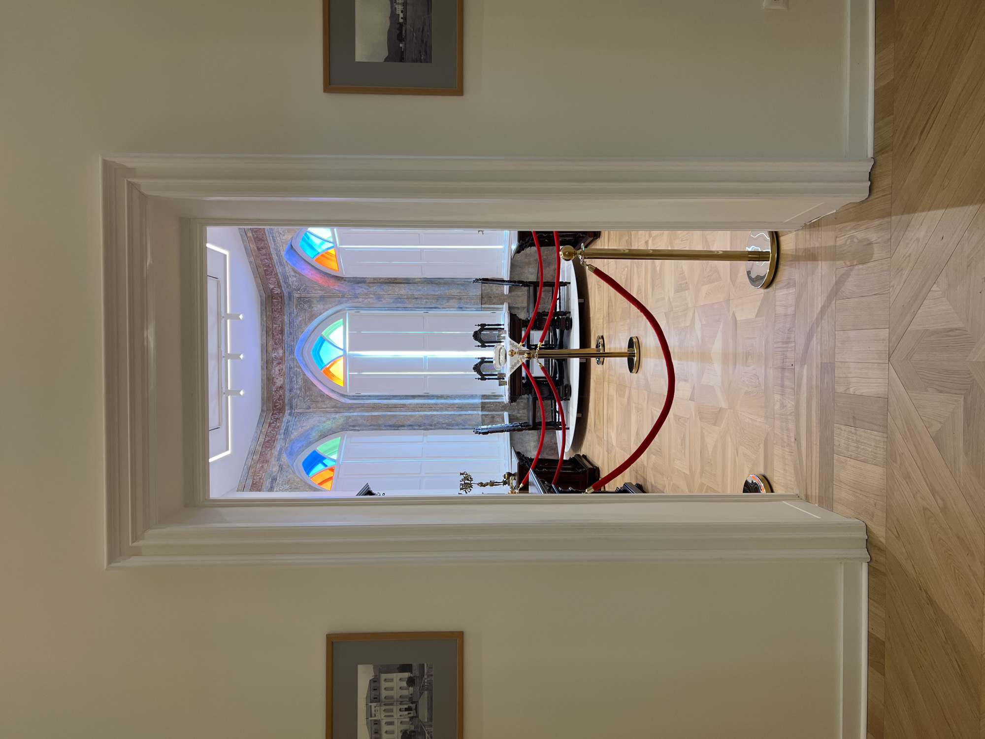 The image shows an interior view of what appears to be a historical building or museum room, possibly located in Bar, Montenegro, as suggested by the context. The scene is viewed through an open doorway with white trim and molding.
In the foreground, a polished wooden floor with a parquet pattern is visible, extending from the hallway into the room. A red velvet rope barrier with gold stanchions is set up to guide visitors, indicating a preserved or exhibited space.
Through the doorway, a room is visible featuring a large oval table with several chairs around it. Behind the table, three tall windows with arched tops are prominent. These windows are adorned with stained glass in vibrant colors, including blue, green, orange, and red, casting colorful light into the room. The upper parts of the walls are decorated with a fresco or mural. White louvered shutters are fitted behind the stained glass windows.
On the walls of the hallway leading to the room, two framed photographs are displayed. The photograph on the left is a black and white image of a large, multi-story building with a garden or courtyard. The photograph on the right is also a black and white image, depicting a building with a hilly landscape in the background.
The room itself contains ornate furniture, including the table and chairs, and what appears to be a candelabra or decorative lamp on a side table. The overall impression is one of historical preservation and an attempt to recreate or showcase a past era.