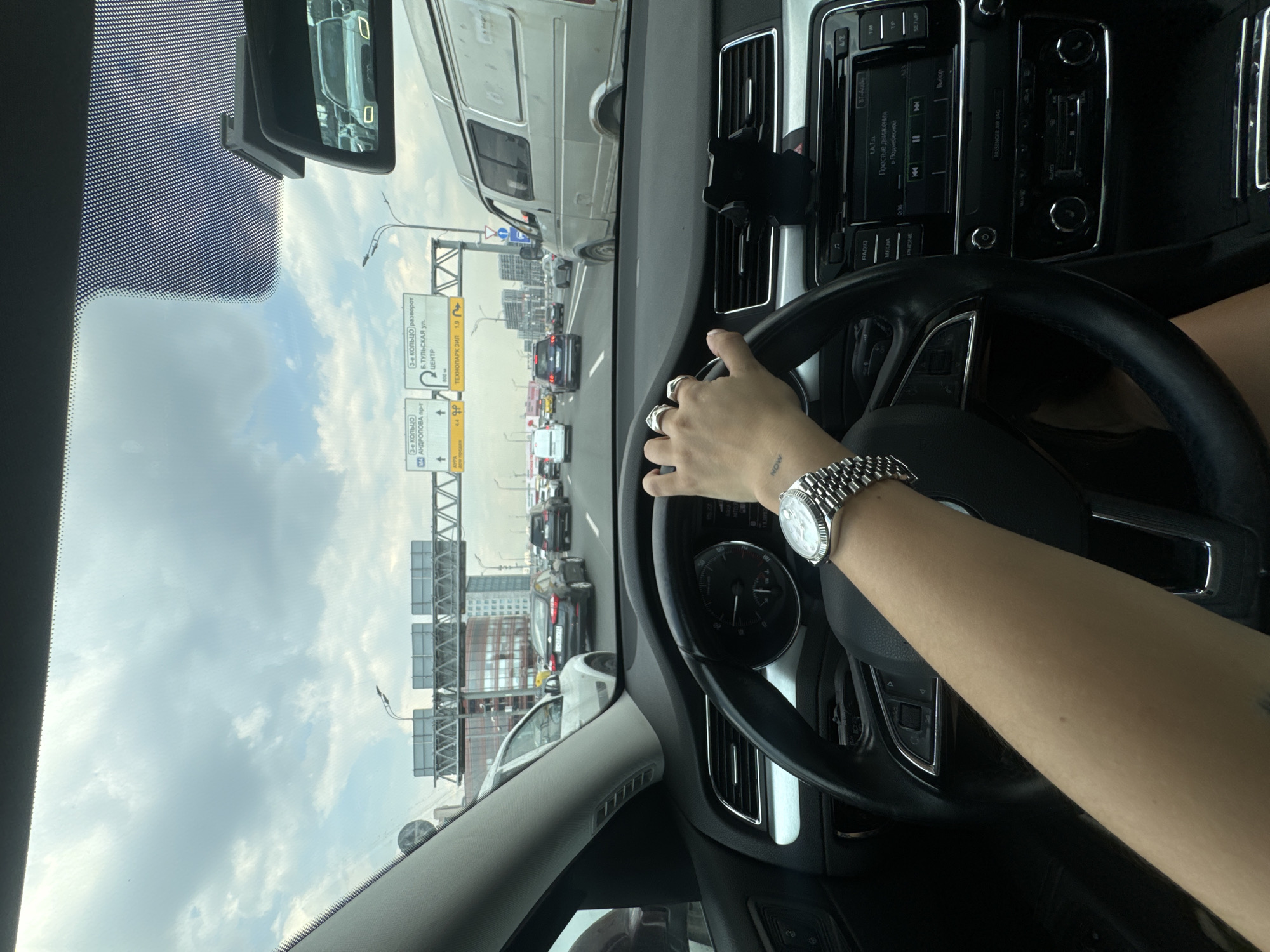 A first-person perspective shot shows a person driving a car on a multi-lane highway in Moscow, Russia, under a cloudy sky. The driver's left hand, adorned with rings and a watch, is on the steering wheel. The car's dashboard is visible, including the radio display showing "Простые движенья" (Simple Movements) and "в Подмосковье" (in the Moscow region). Road signs indicate directions to "3-е КОЛЬЦО" (3rd Ring Road) and "ул. ТУЛЬСКАЯ" (Tulskaya Street), suggesting the location is near or on a major artery within or leading out of the city. Other vehicles, including cars and a van, are present on the highway, indicating moderate traffic. Buildings and infrastructure typical of a city environment are visible in the background.