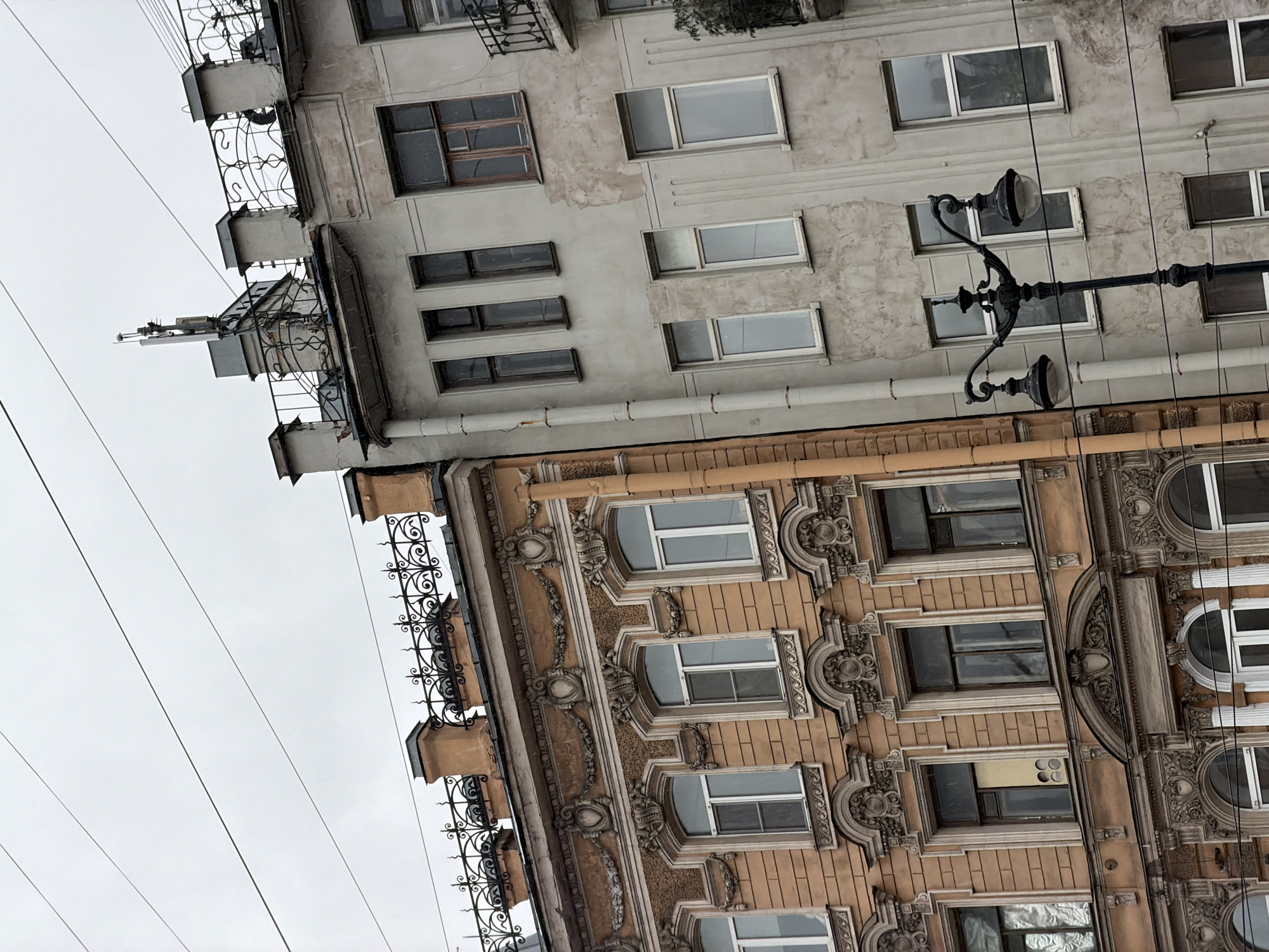 A tilted low-angle shot captures two buildings in Saint Petersburg, Russia, under an overcast sky. The upper building is painted in muted grey tones with rectangular windows of varying sizes. It features small iron balconies with decorative railings. Above the balconies is an antenna. The lower building is a warm tan or orange color, with more elaborately decorated windows and classical architectural details. A black lamppost with two round lamps extends from the upper building downwards. Power lines crisscross the sky in the upper left corner.
