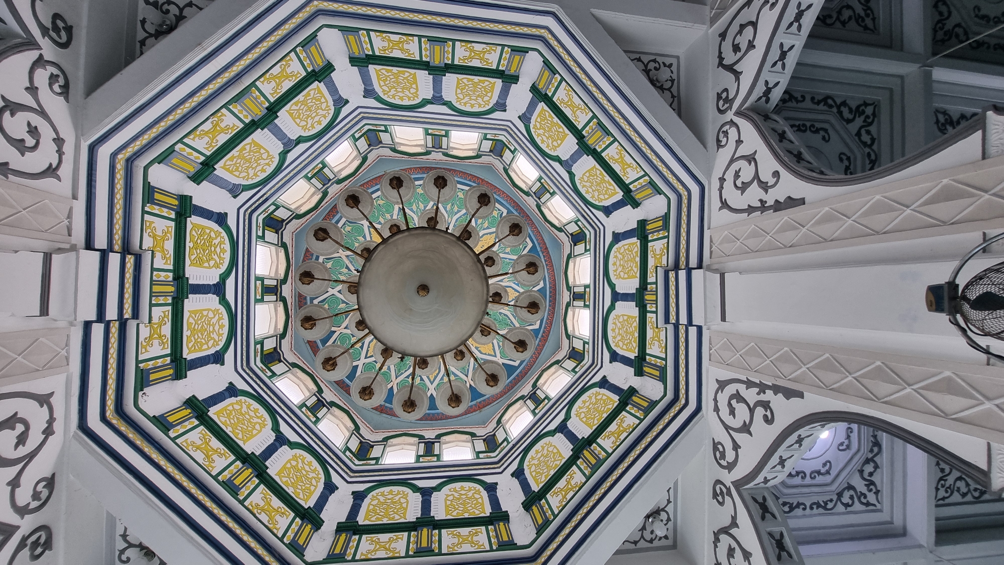 An interior shot of a domed ceiling in the Keudekruenggeukueh, Indonesia, features a large, ornate chandelier suspended from the center. The ceiling is octagonal and decorated with intricate patterns in blue, yellow, green, and white. Each facet of the octagon has painted designs resembling stylized floral motifs and geometric shapes. The chandelier itself has a circular central fixture with multiple arms, each holding a glass light cover. Below the main dome, architectural elements of the building are visible, including arched doorways with decorative grey scrollwork and diamond-patterned white columns. A small, dark electric fan is positioned on a lower ledge, partially obscuring one of the architectural columns. The lighting suggests it is daytime, with light filtering in from windows or openings within the dome structure.