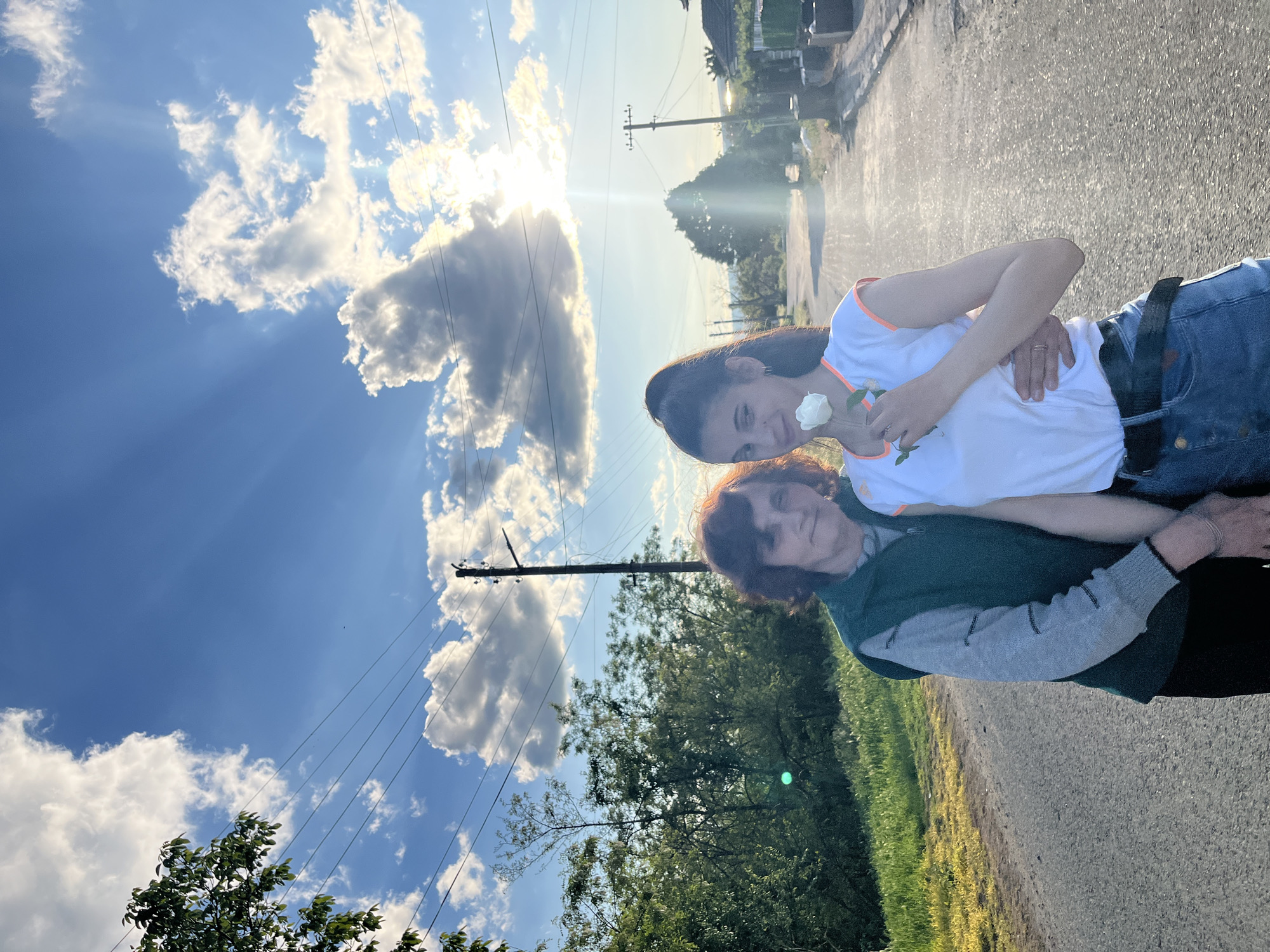 The image depicts two females on a paved road in Lozarevo, Bulgaria. One female is standing and holding a white rose. She wears a white t-shirt with orange trim and blue jeans. Her arm is around the shoulders of another shorter female. The shorter female is wearing a dark green vest over a grey sweater. They are positioned on a road with vegetation and a grassy area on one side. Overhead power lines are visible. In the background, there are buildings and a sky with clouds and the sun.