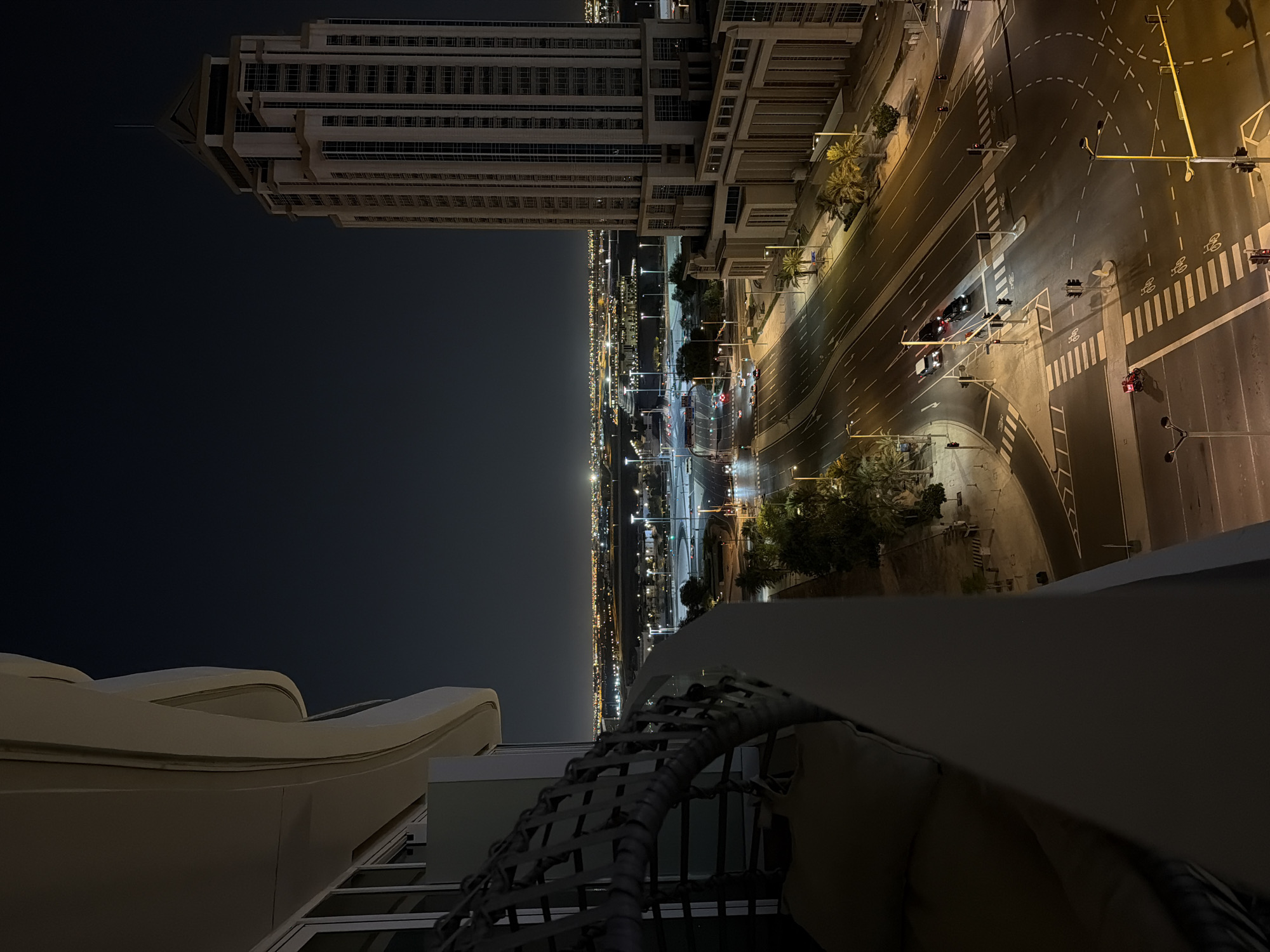 This is a high-angle, nighttime photograph overlooking a city scene. In the foreground, on the left, a portion of a balcony or terrace is visible, featuring a hanging chair with cushions. To the right, a tall, modern skyscraper dominates the mid-ground, its windows illuminated. A multi-lane road is visible in front of the skyscraper, with several cars driving and parked. Streetlights illuminate the road and sidewalks. In the background, a sprawling cityscape stretches to the horizon, dotted with numerous lights, suggesting a dense urban environment. The sky is dark and clear, indicating nighttime.