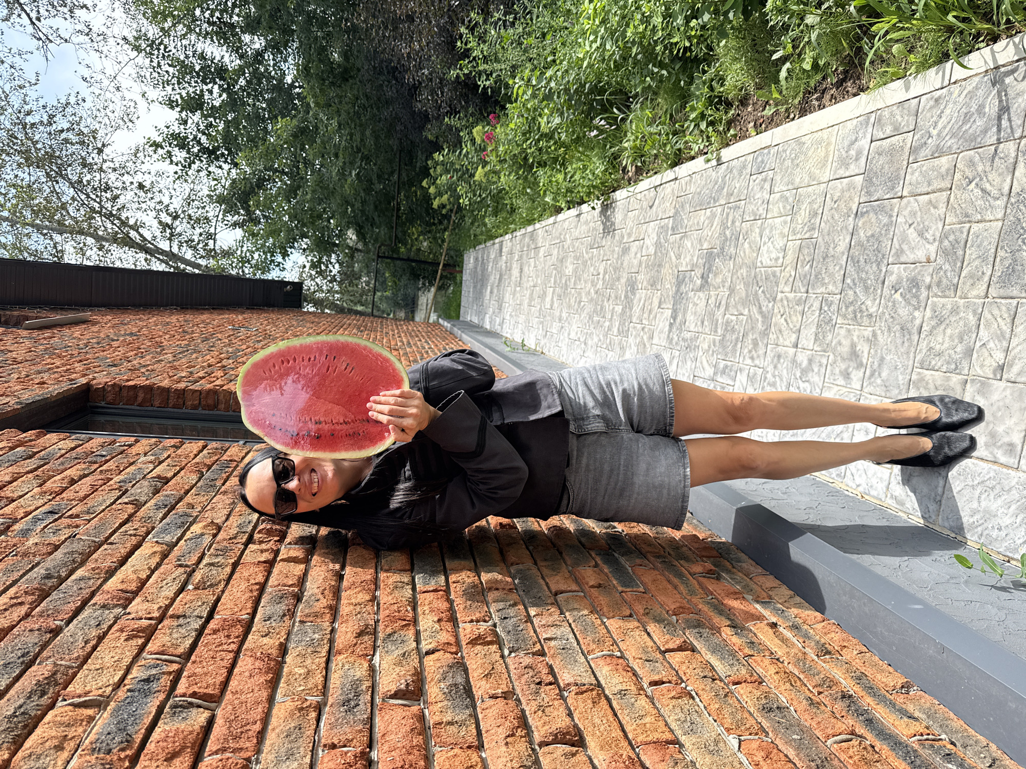 A woman stands outdoors leaning against a red brick wall, holding a slice of watermelon. She wears sunglasses, a dark jacket, grey denim shorts, and black heeled shoes. The sidewalk in front of her is paved with light-colored interlocking stones, bordered by lush green foliage. The scene is set in a bright, sunny environment, likely a residential area or garden in Chaikovsky, Russia.
