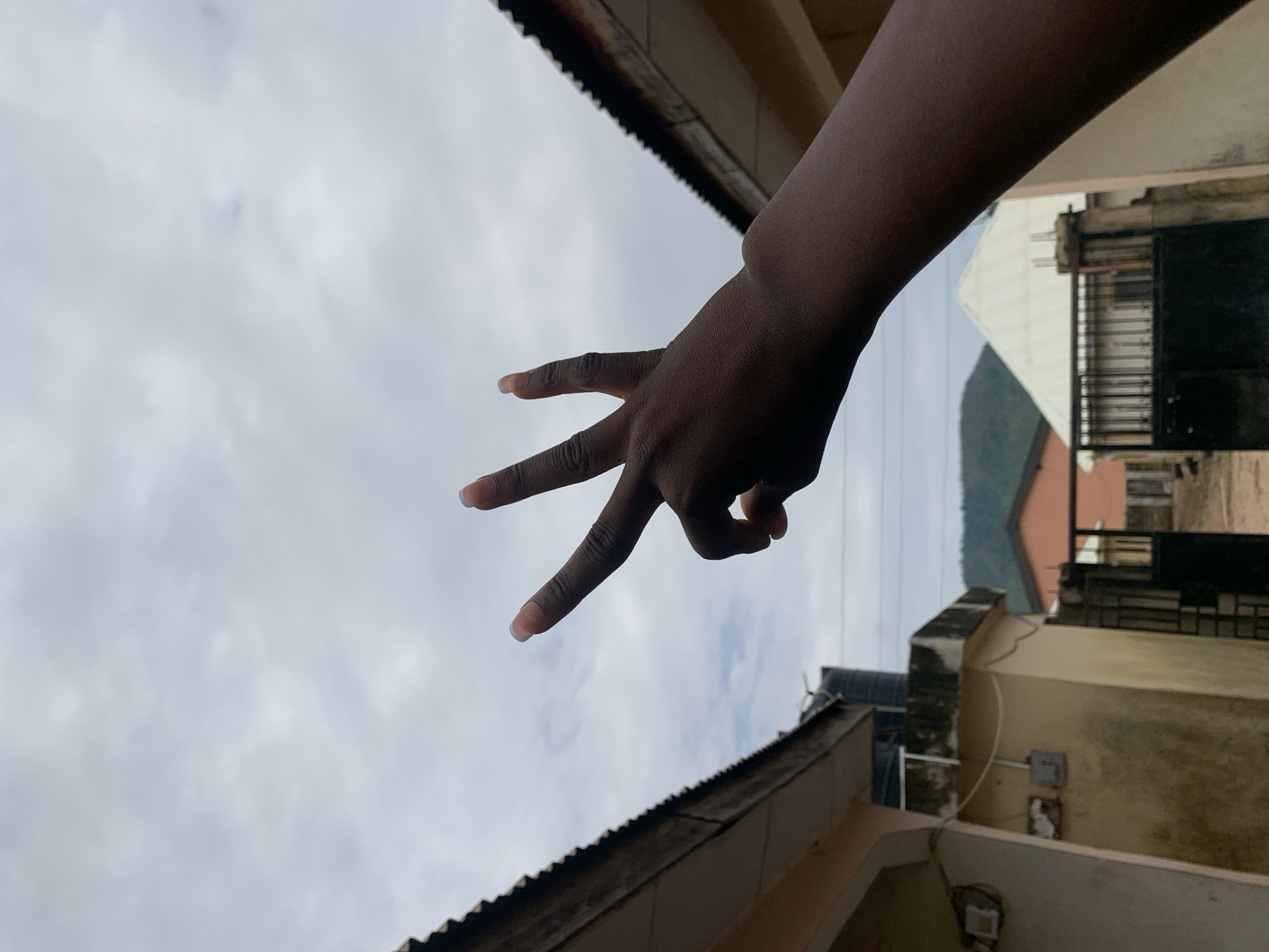 An arm is extended at an angle from the upper right of the frame. The hand is displaying three fingers, with the index, middle and ring fingers extended and the pinky and thumb tucked into the palm. The person has brown skin and manicured fingernails. The backdrop consists of a sky with a dense overcast of grey clouds in the background and the facades of adjacent buildings. There is a building with a brown, rusty corrugated metal roof on the left. The other buildings are a cream color with dark grey metal security bars on the windows. A mountain in the background is seen in the right distance. This image was taken in Lokoja, Nigeria.