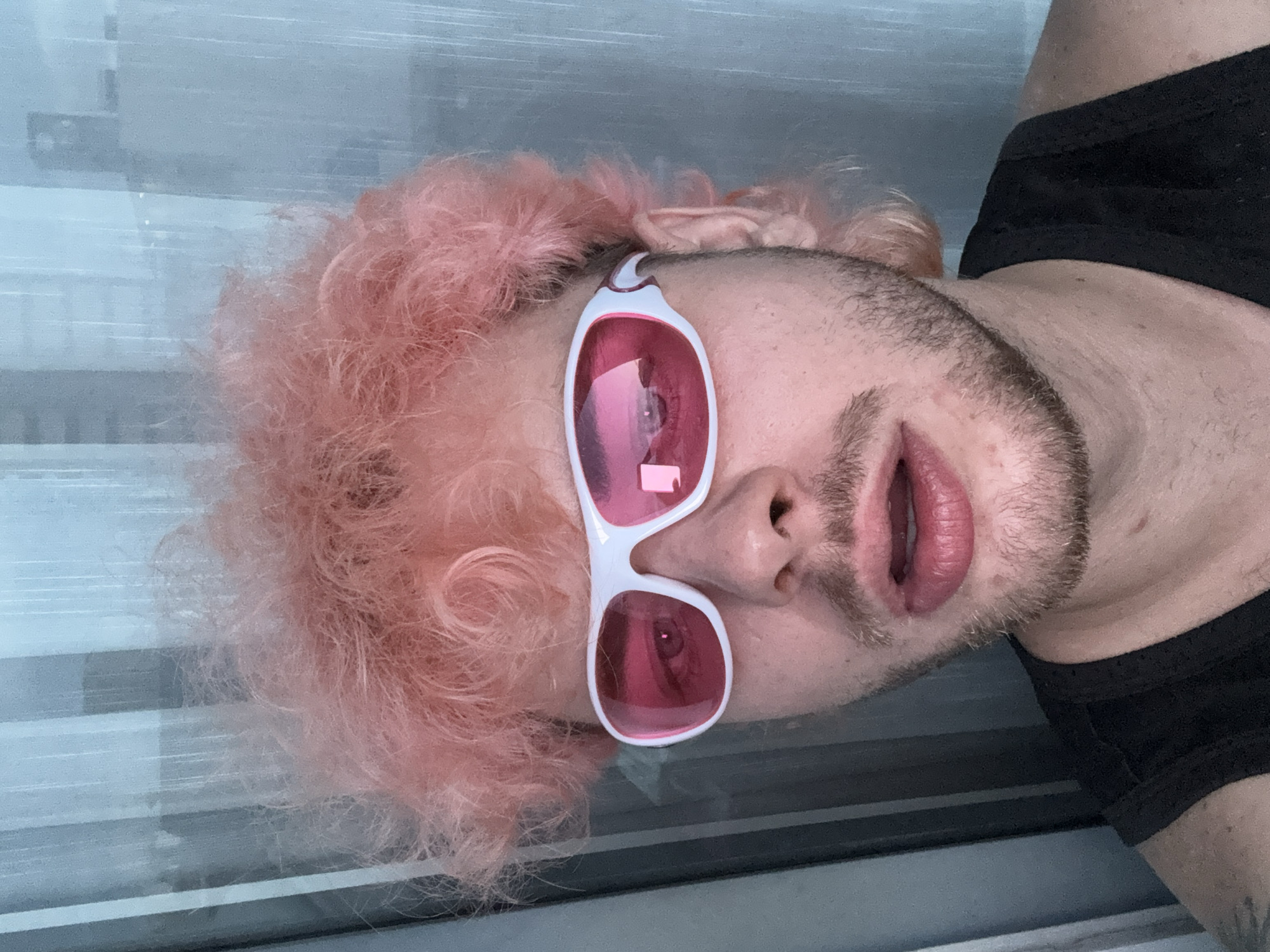 The image shows a close-up, head-and-shoulders portrait of a person with light pink, voluminous, curly hair. They are wearing white-framed, pink-lensed wraparound sunglasses and a black tank top. The person's mouth is slightly open, revealing their teeth. Their skin has a light complexion, and they have facial hair. The background consists of sheer, vertical blinds that are translucent and allow some light to pass through, creating a softly blurred effect. No specific location in Đà Nẵng, Vietnam can be identified from the image.