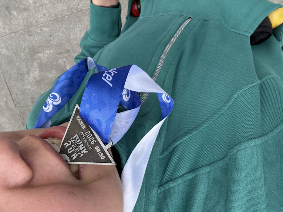 A person wearing a green zip-up hoodie is lying down, partially visible. A "Vania 2026 Think Feel Run" medal dangles from their mouth, held by the teeth. The medal is attached to a blue and white ribbon with circular patterns. The person's face is in the bottom left, showing a slight smirk. The background shows what appears to be concrete.