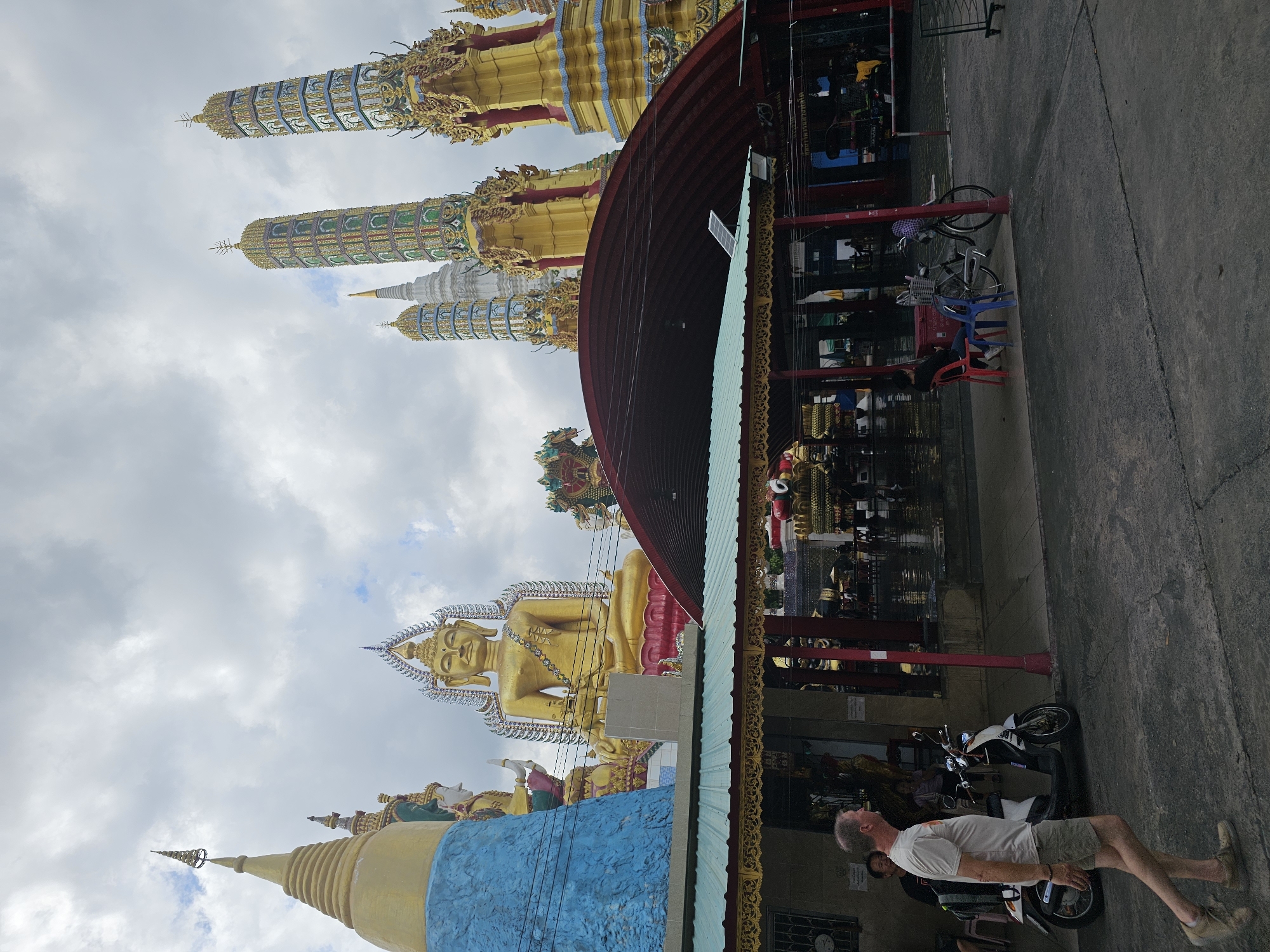 In Bangkok, Thailand, a man walks past a Buddhist temple. The temple features several tall, ornate stupas, a large golden Buddha statue, and intricate architectural details. The sky is cloudy, and the ground is paved. Several people are visible inside the temple complex, some seated and others walking. A scooter and a bicycle are parked near the entrance. The scene appears to be a typical day at a religious site, with a mix of religious structures and everyday objects.